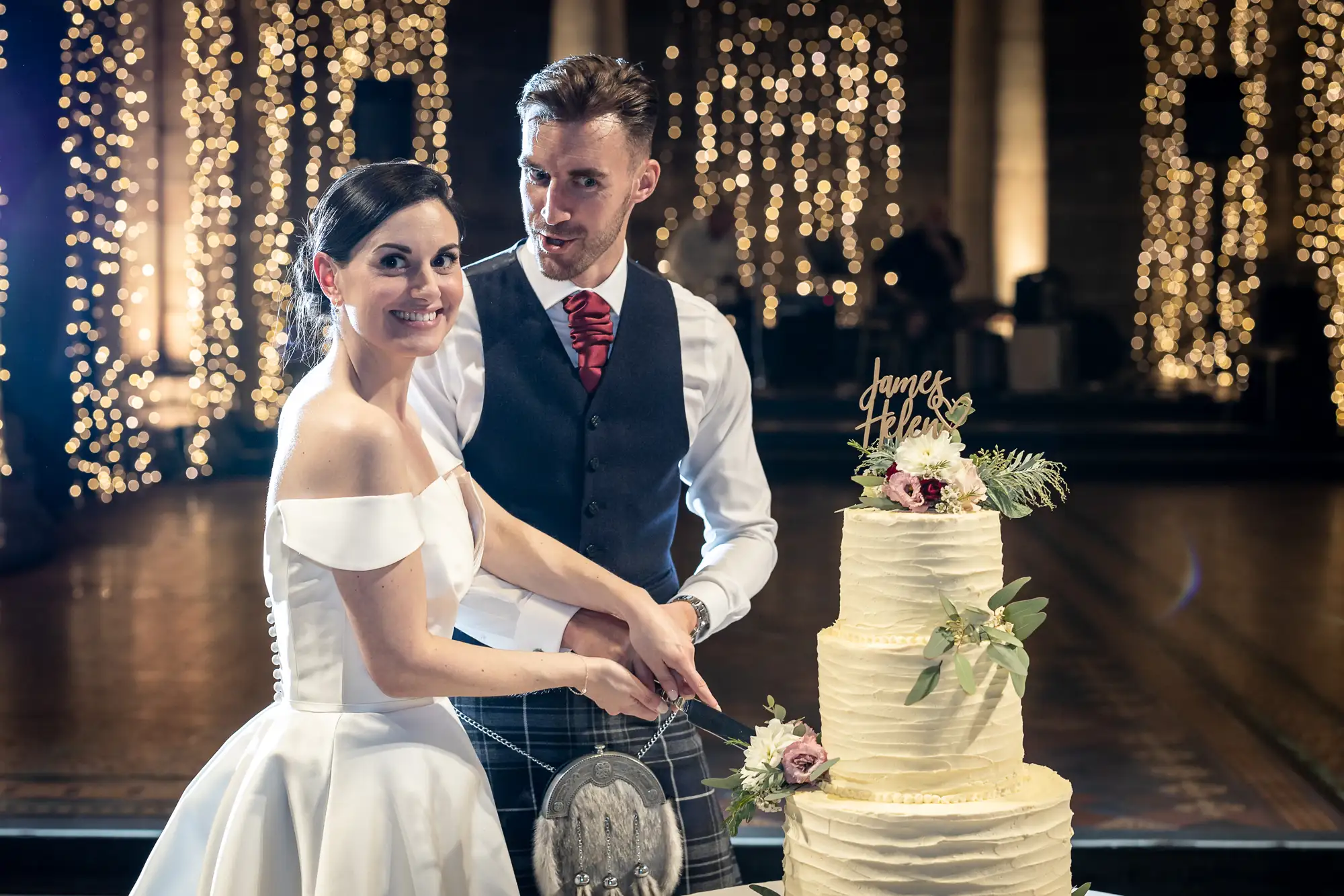 A bride and groom cut a wedding cake together in a hall decorated with fairy lights, looking at the camera with smiles.