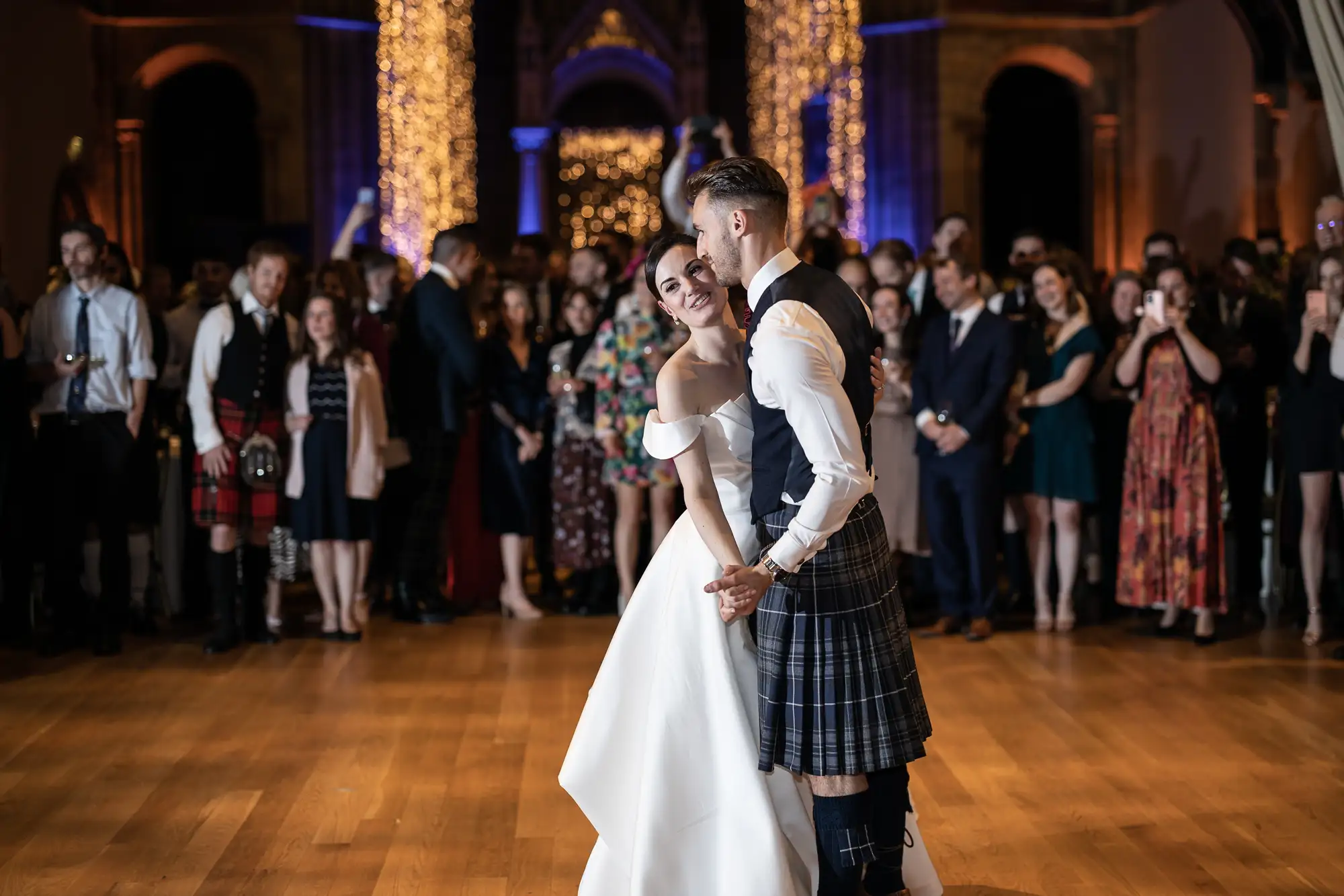 A bride in a white gown and a groom in a kilt share a dance, surrounded by guests in a warmly lit hall.