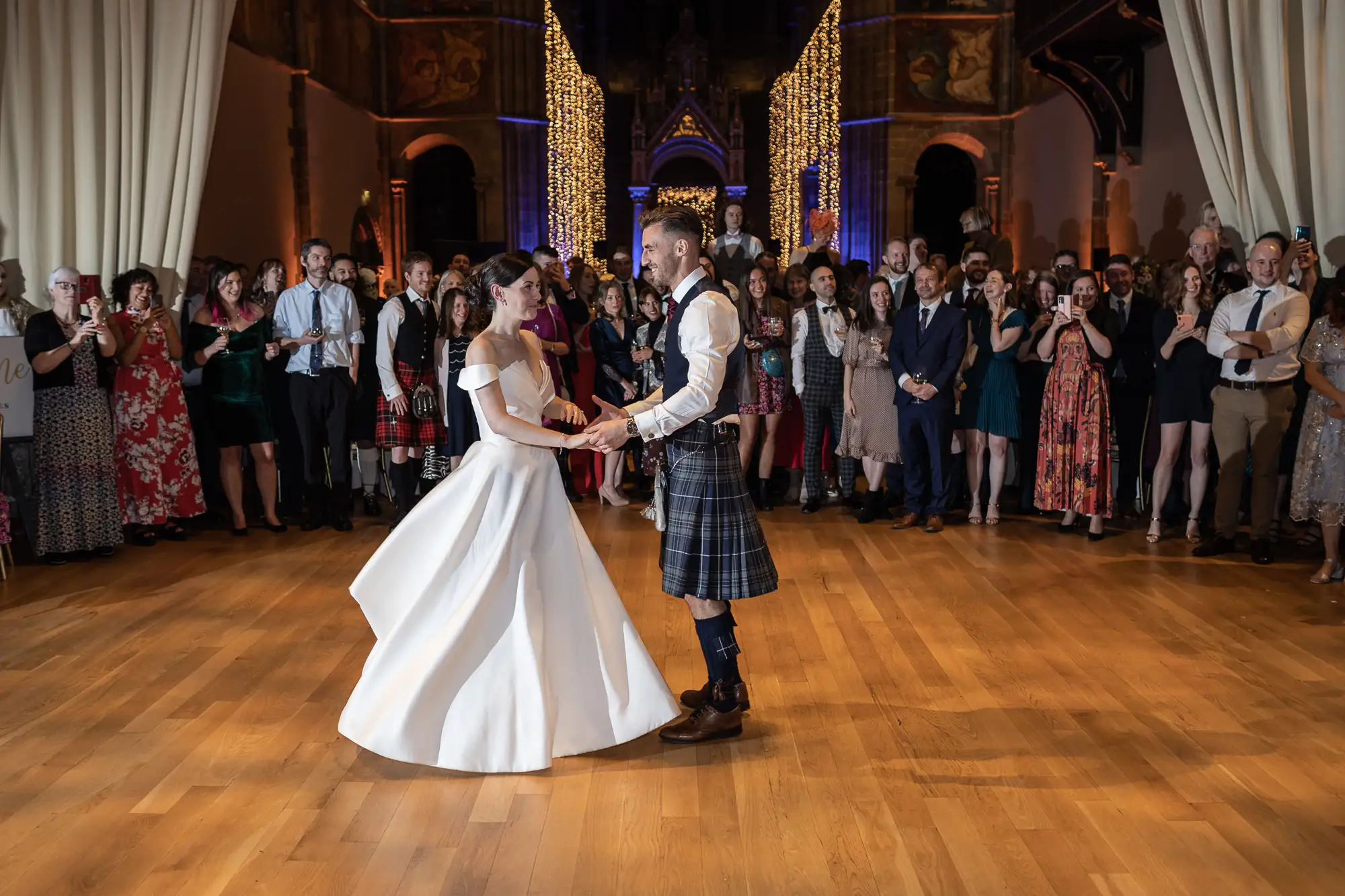 A bride in a white dress and a groom in a kilt dance in the center of a ballroom, surrounded by guests watching and clapping.