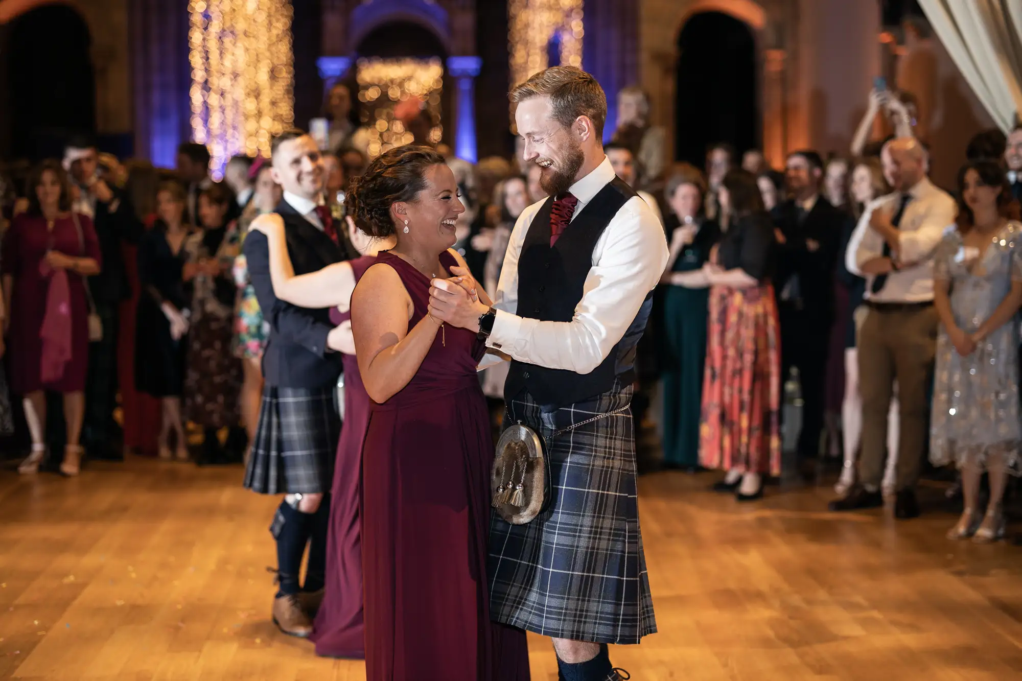 A man in a kilt and a woman in a burgundy dress share a dance at a formal event, surrounded by onlookers in a well-lit, ornate ballroom.