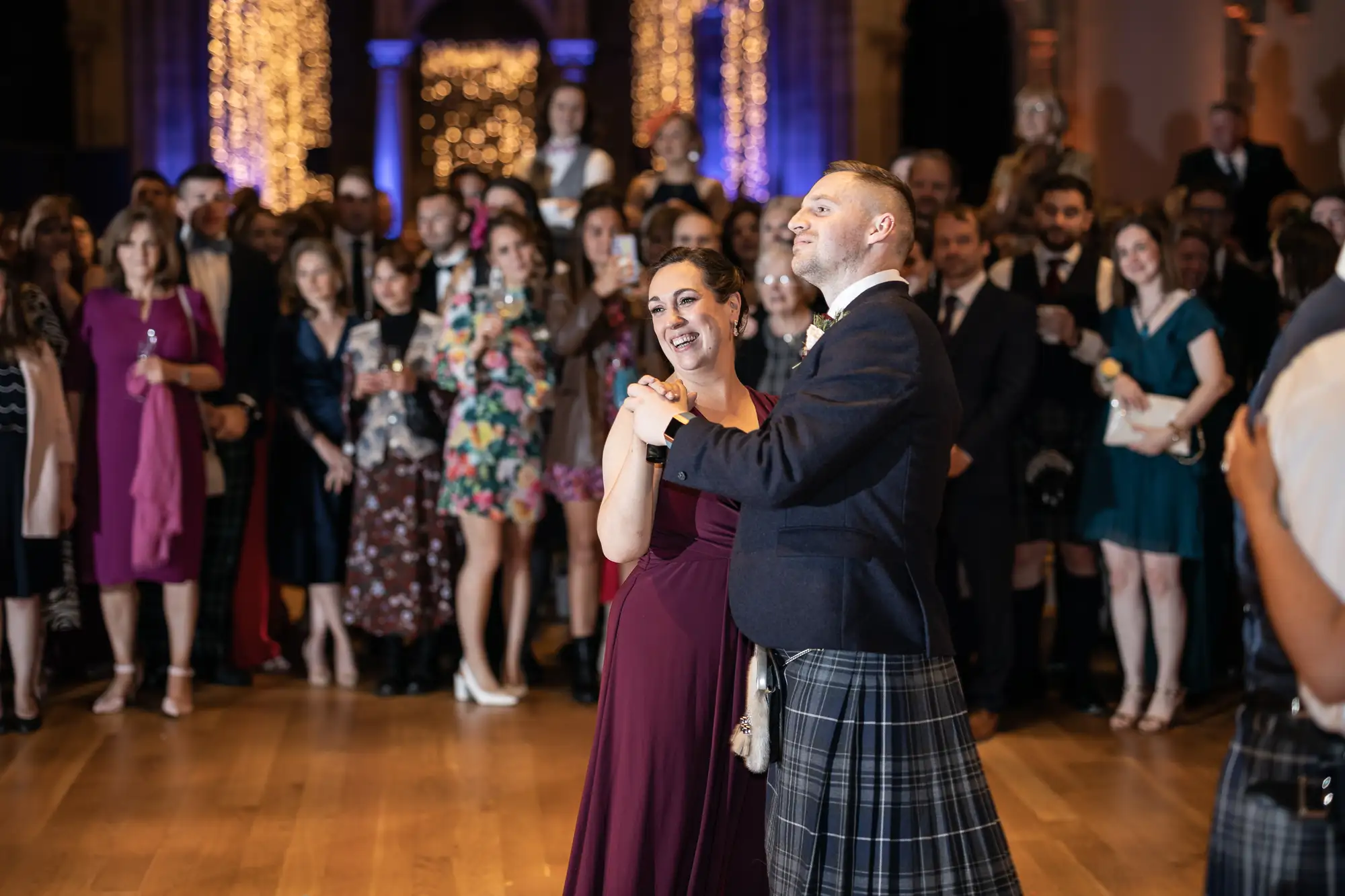A couple joyfully dances in front of a crowd at a formal event, the man wearing a kilt and the woman in a burgundy dress.