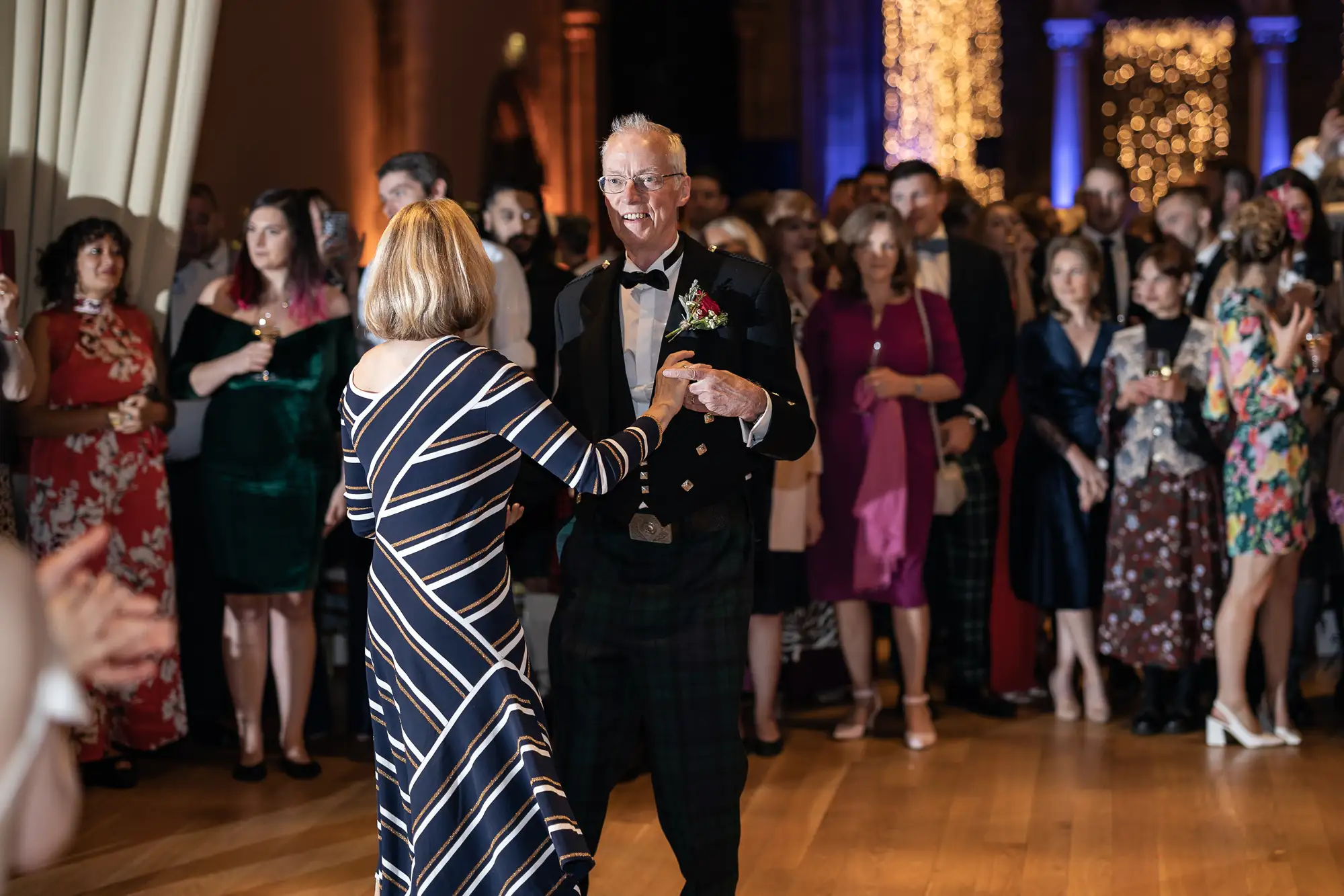 An elderly couple dances joyfully at a formal event, surrounded by onlookers in a warmly lit hall. the man wears a traditional kilt, and the woman dons a striped dress.