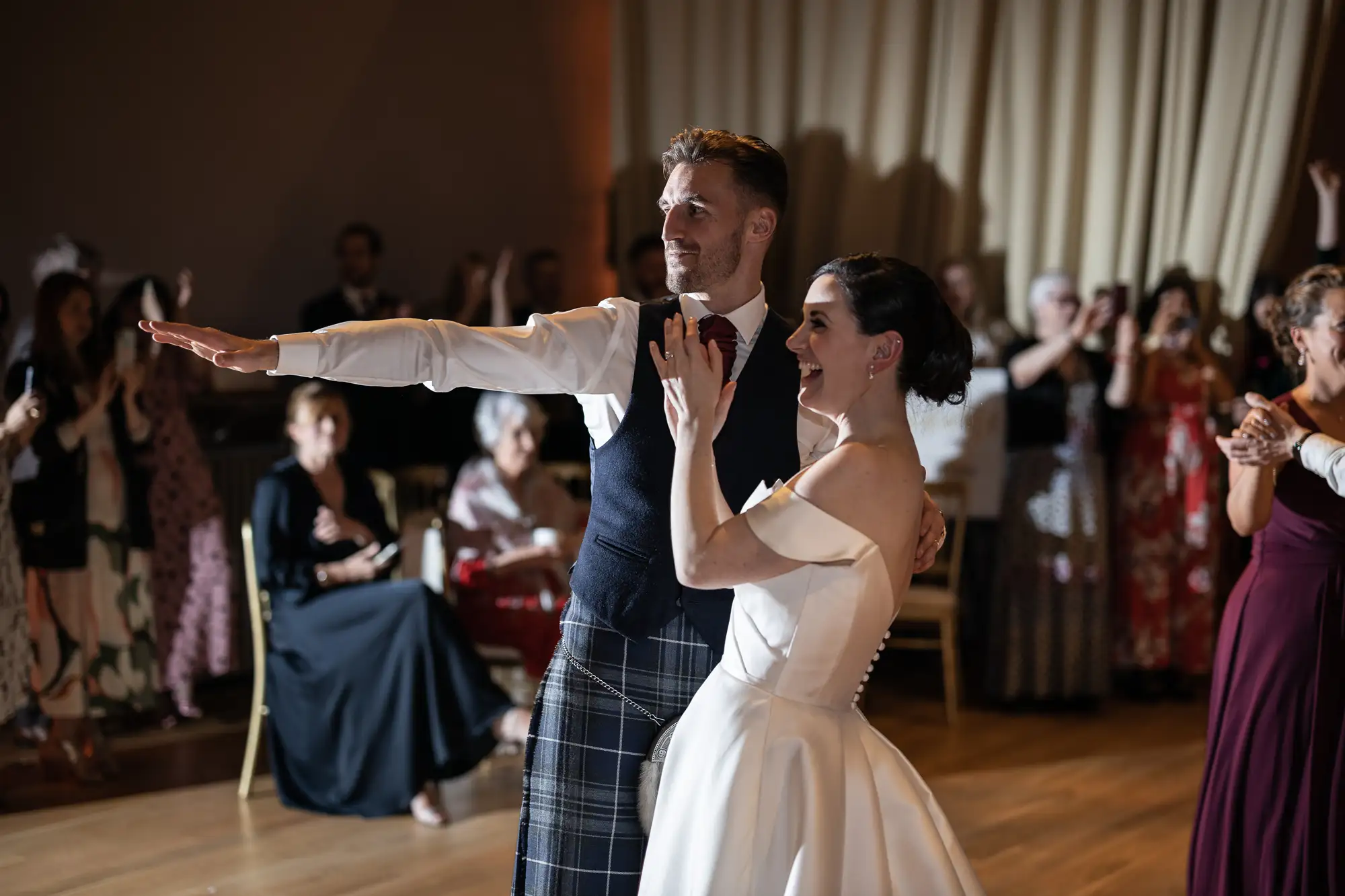 A bride and groom joyfully dancing in front of wedding guests, the bride in a white dress and the groom in a kilt.