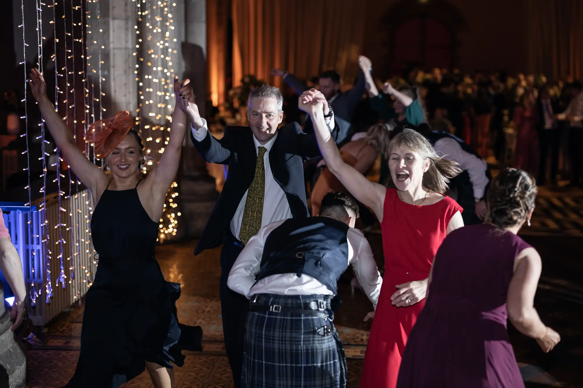 Guests happily dancing at a formal event, with lights hanging in the background and one man in a kilt leading a cheerful dance.