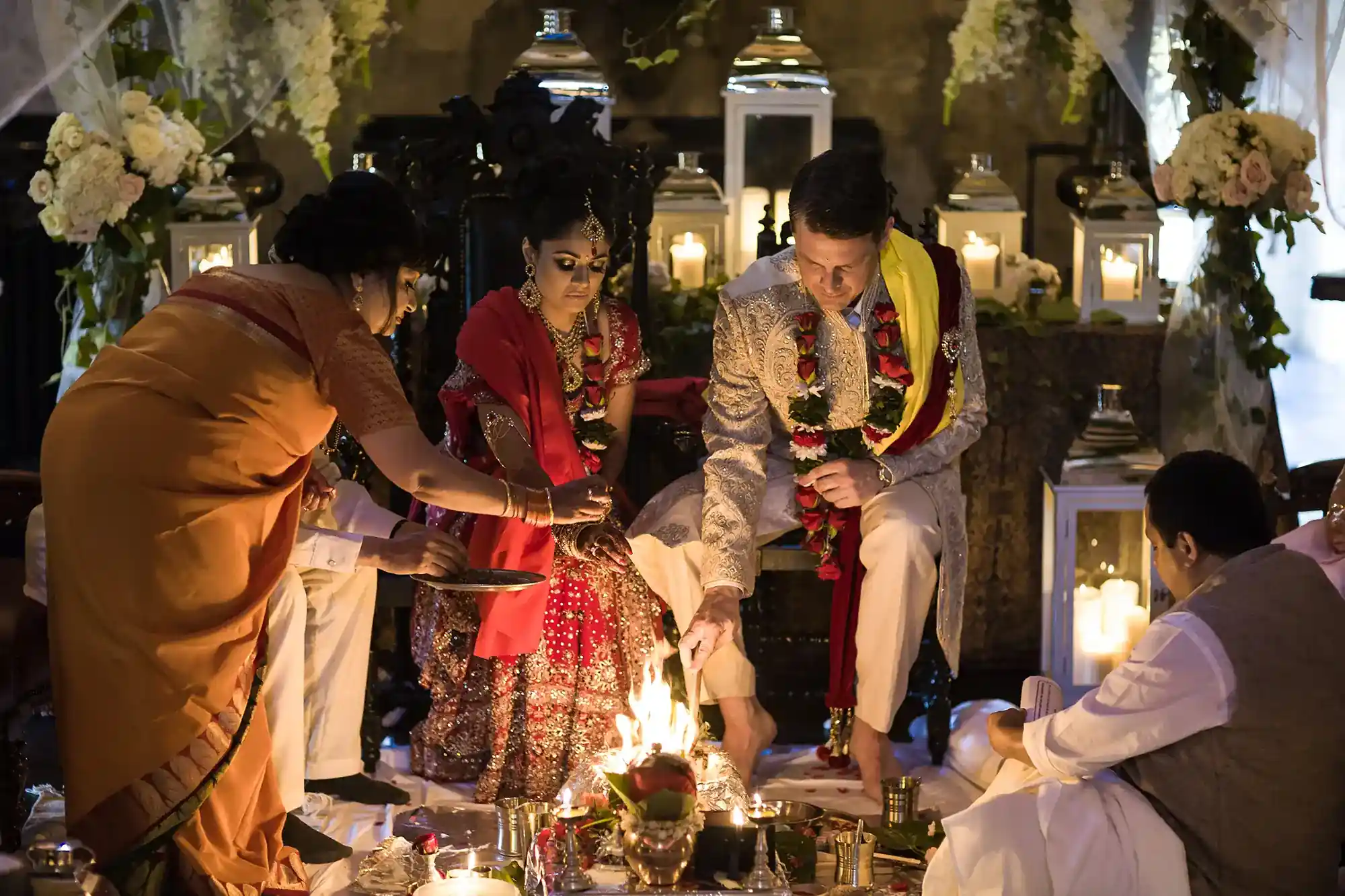 A bride and groom sit beside a sacred fire during a Hindu wedding ceremony, surrounded by ritual items and attendees performing traditional rituals.