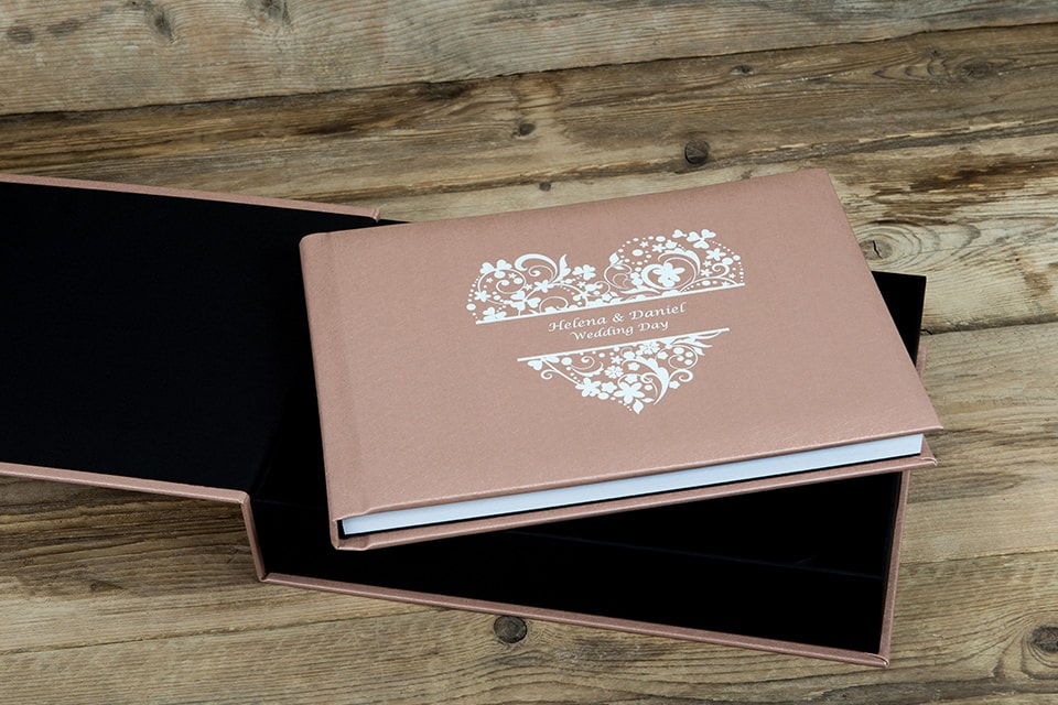 Wedding album cover and box example: A coffee coloured wedding photo album with white floral heart design and the names "Helena & Daniel" alongside "Wedding Day" is partially open on a wooden surface.