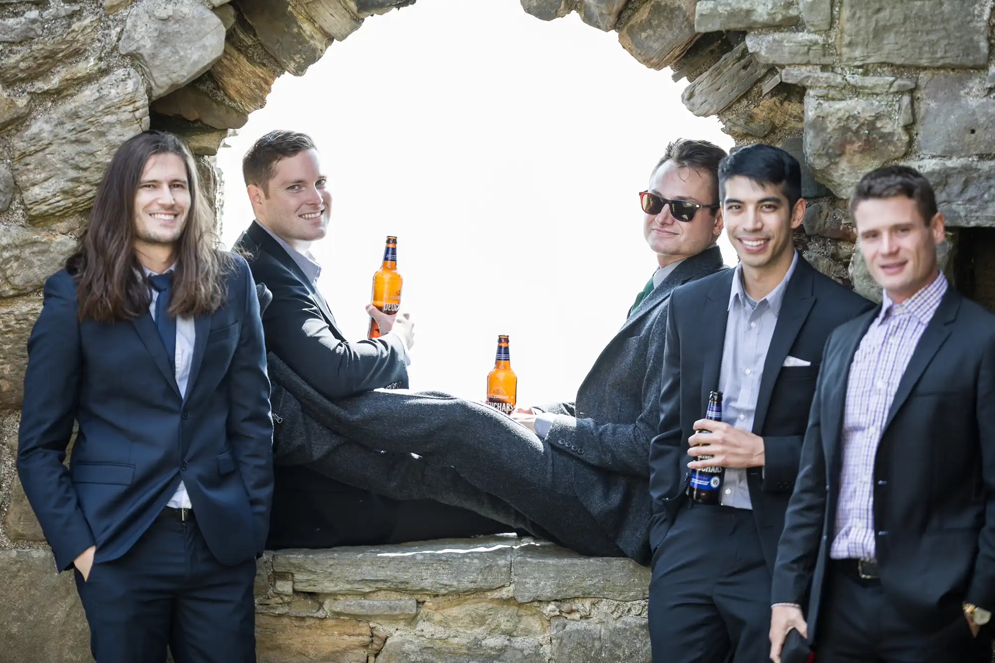 Five men in suits stand and sit by a stone archway, holding bottles of beer and smiling.