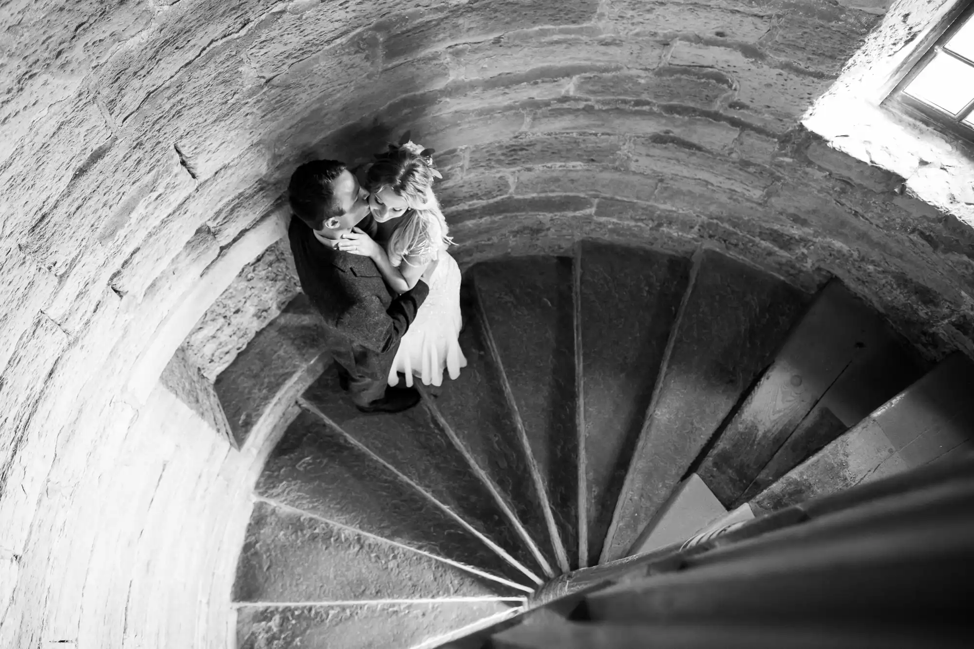 A black and white photo shows a newlywed couple embracing on a spiral staircase, viewed from above. The staircase is made of stone, and sunlight streams in from a small window.