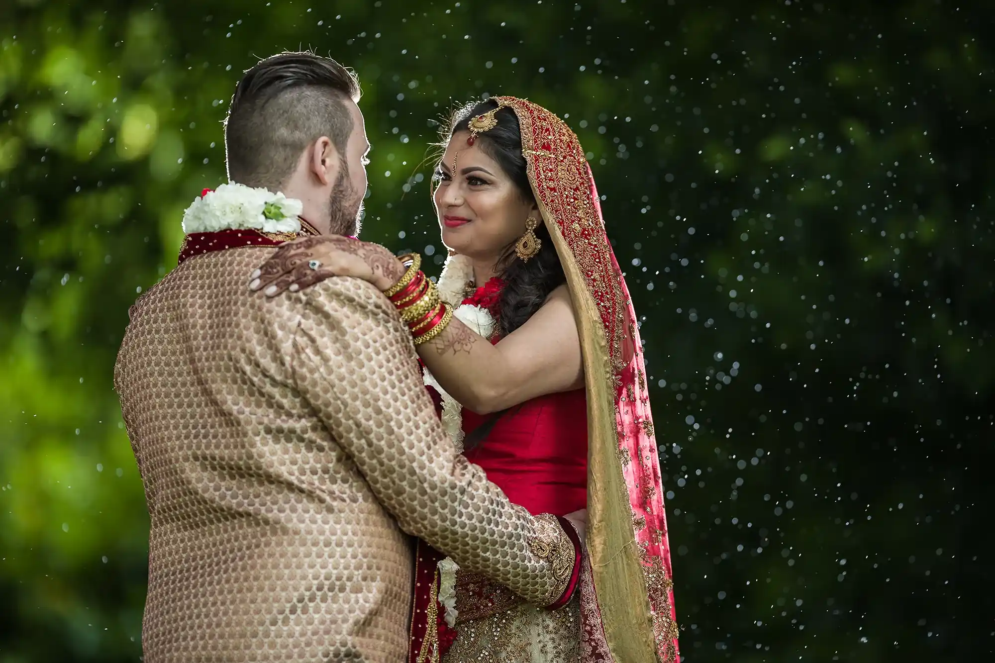 A bride in a red saree and groom in a beige sherwani share a moment under a sprinkle of raindrops, surrounded by greenery.