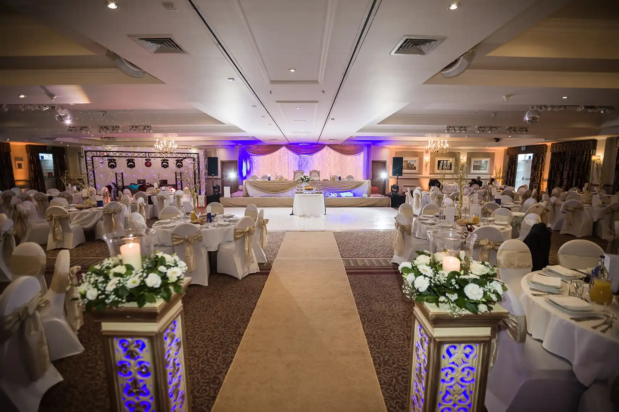A well-lit wedding reception hall with round tables set with white linens, centered aisle leading to main table under purple lights.