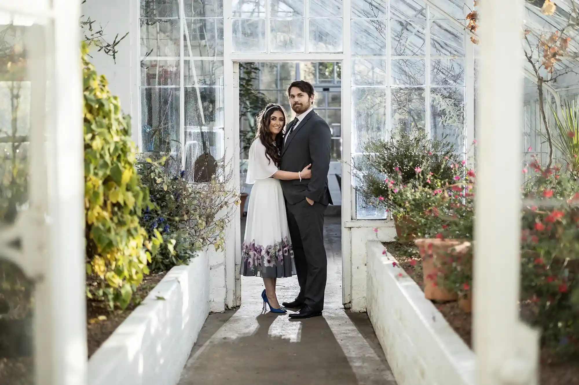 A couple embraces in a sunlit greenhouse, surrounded by lush plants, with the man in a gray suit and the woman in a floral dress.