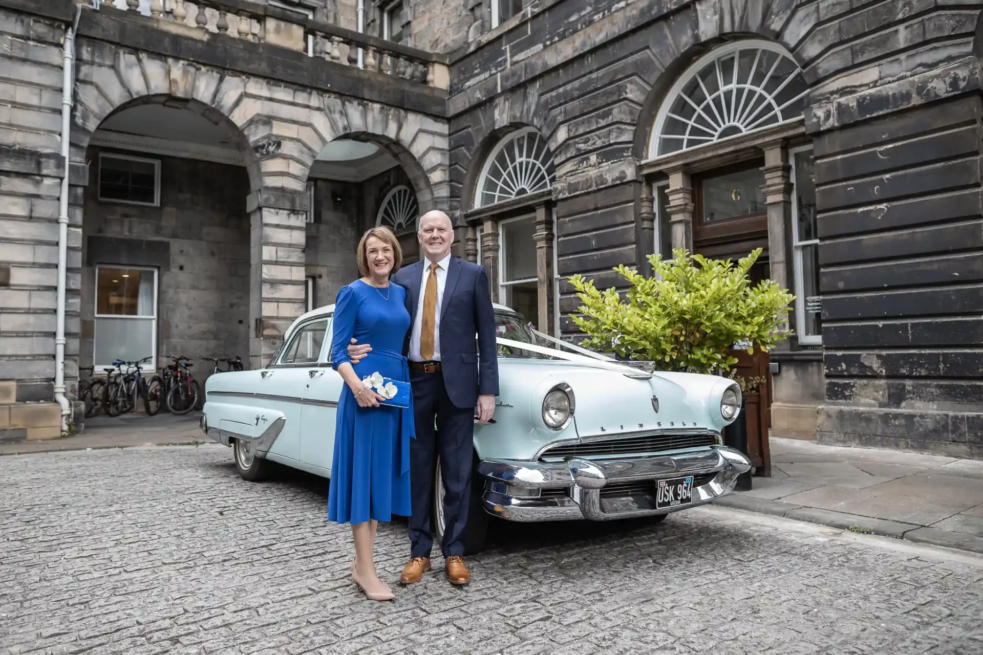 A couple stands in front of a vintage blue car parked by a historic building.