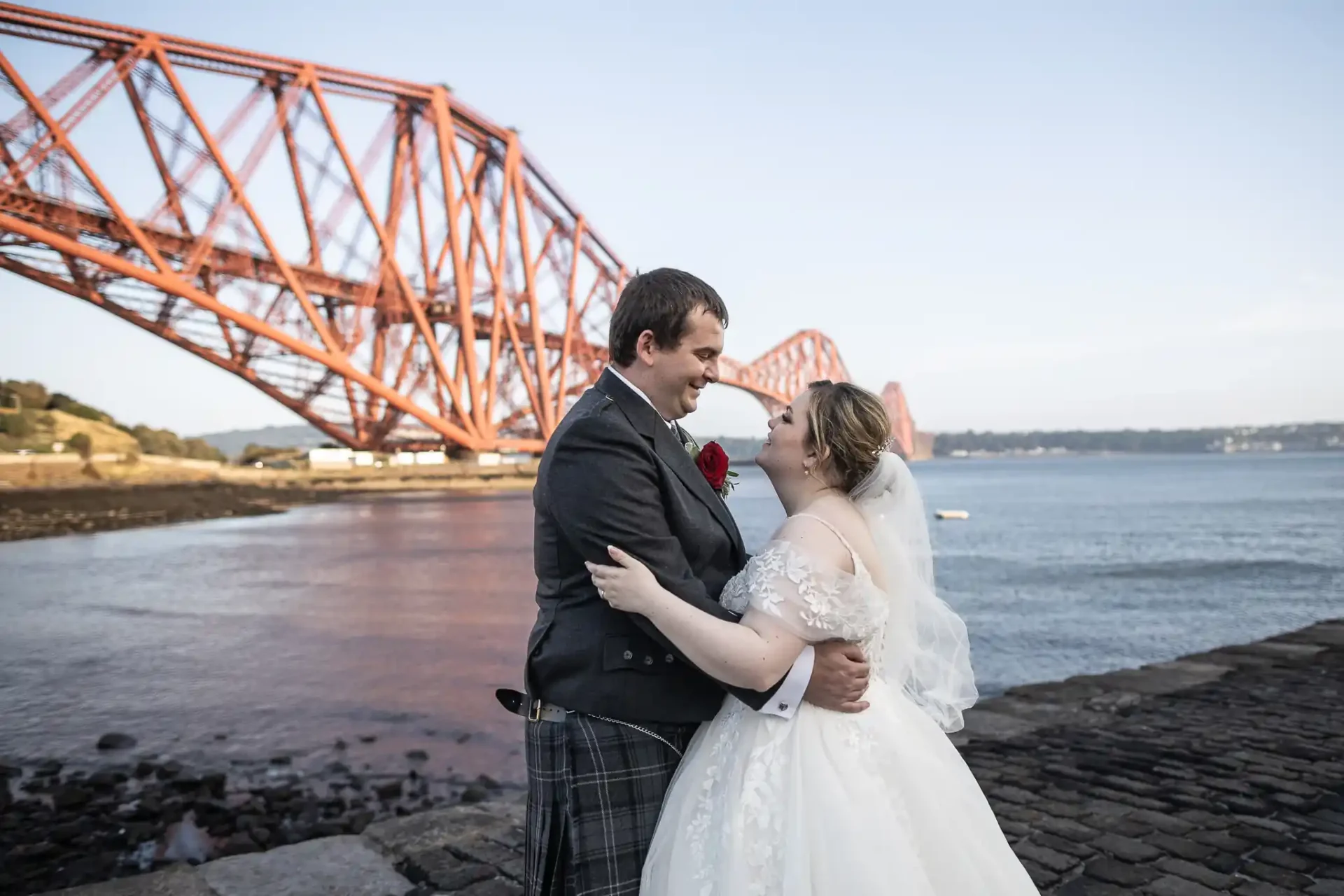 A bride and groom embrace near a large red bridge by the water, with the bride in a white gown and the groom in a suit with a kilt.