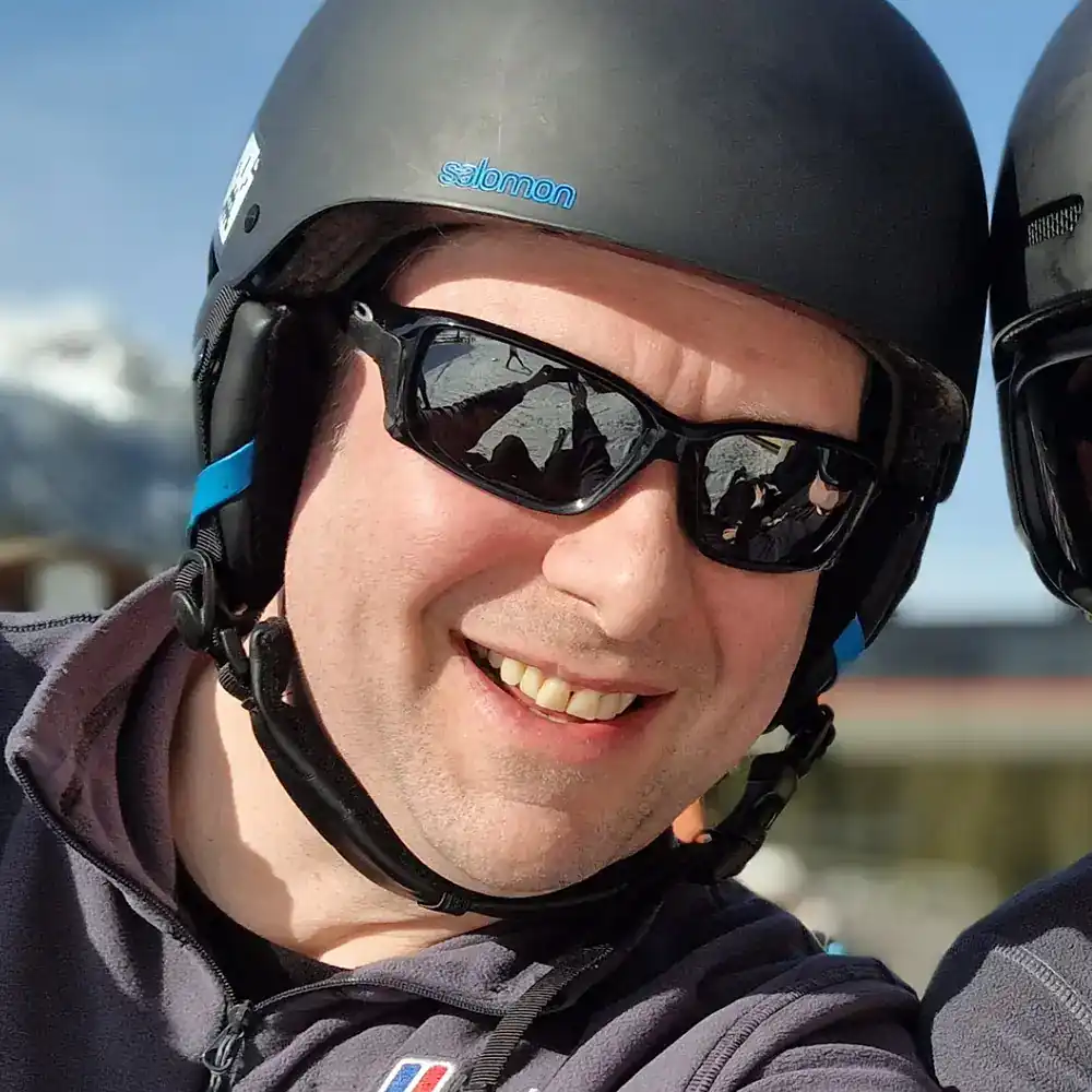 A person wearing sunglasses and a black helmet is smiling at the camera with snowy mountains in the background.