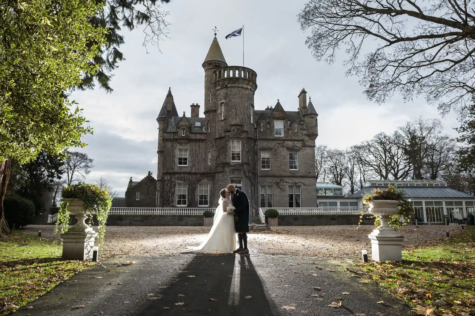 A couple dressed in wedding attire stands in front of a large, historic castle with a flag on top. Trees and greenery surround the area, and the sun casts long shadows on the ground.