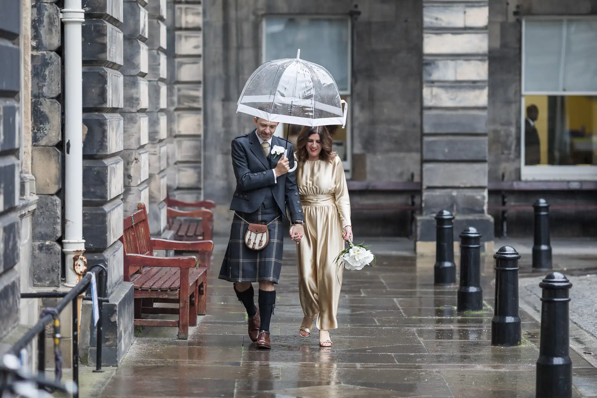 A couple walks under a clear umbrella on a rainy day, with the man wearing a traditional kilt and the woman in a gold dress holding a bouquet. They are walking on a wet stone pathway near a building.