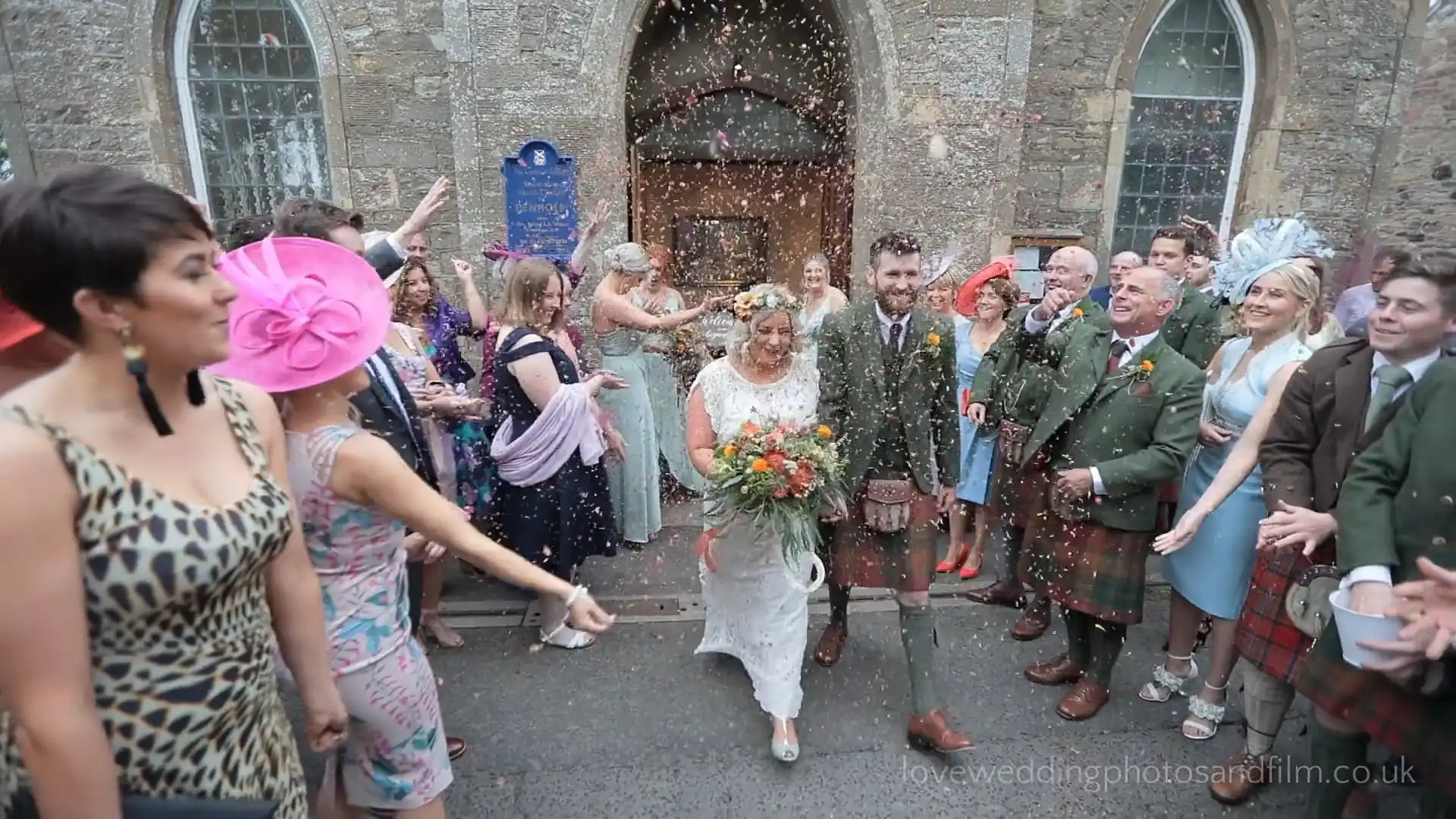 A bride and groom exit a stone church while guests dressed in formal attire throw confetti and celebrate around them. Scottish Borders Wedding Video - Kerry and Liam.