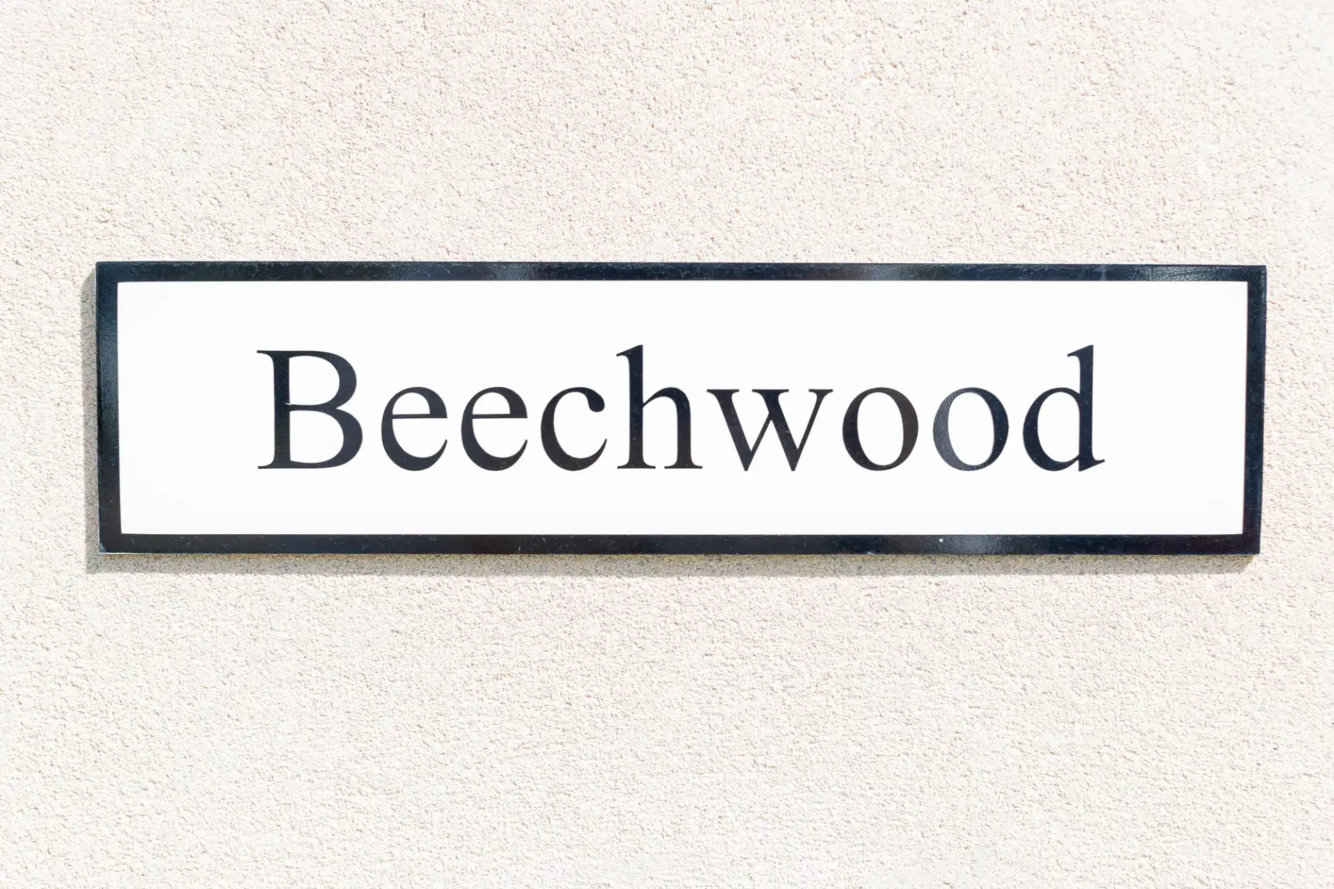 A rectangular sign with the word "beechwood" in black letters, centered on a white background, mounted on a textured beige wall.
