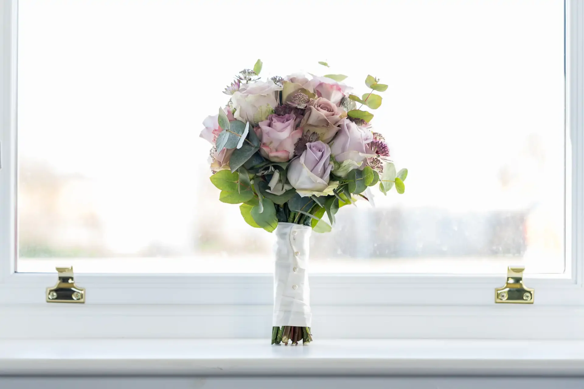 A bridal bouquet with pale pink roses and white flowers wrapped in a ribbon, set against a bright window.