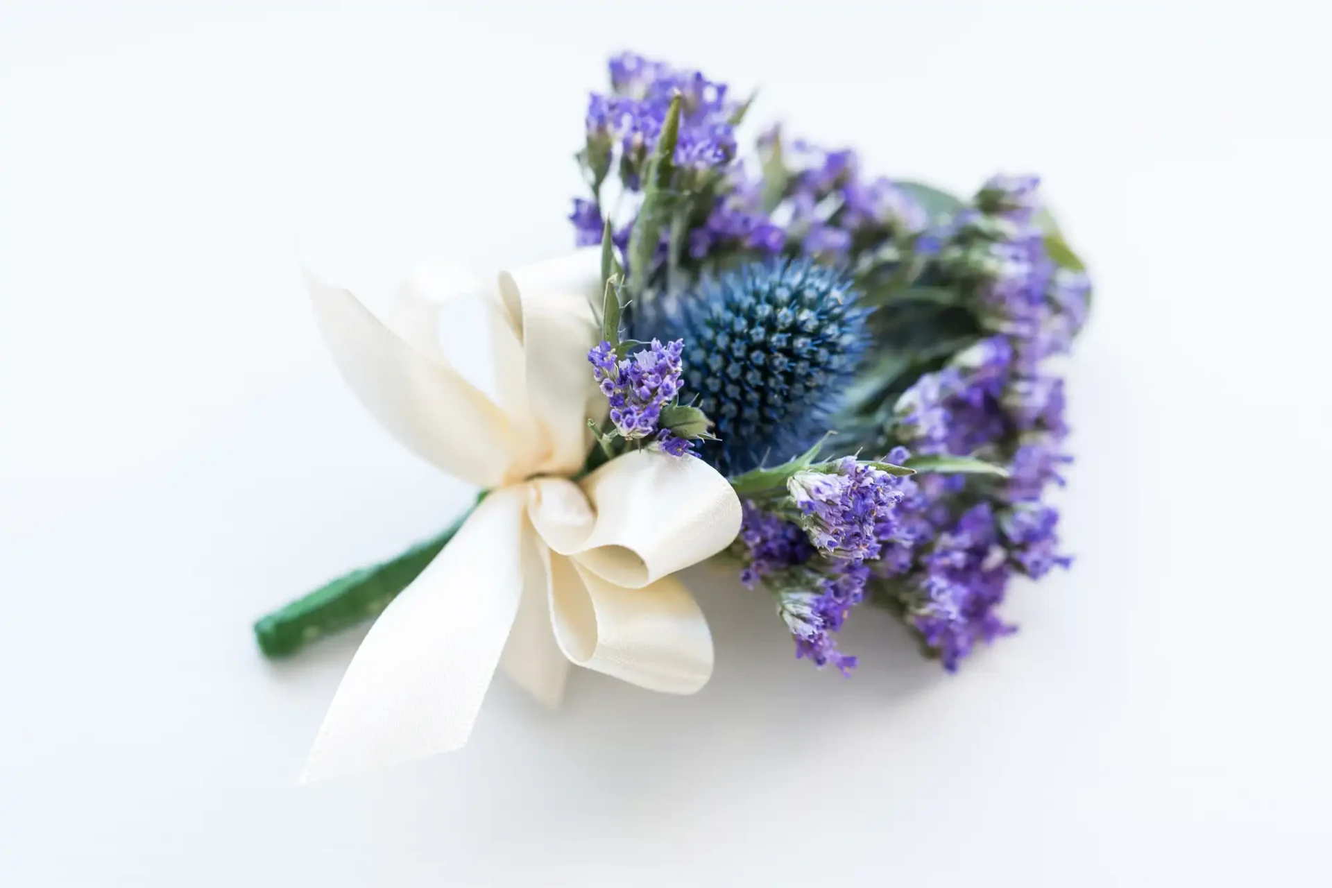 A delicate boutonniere featuring a blue thistle and purple flowers, accented with a large white ribbon, against a white background.