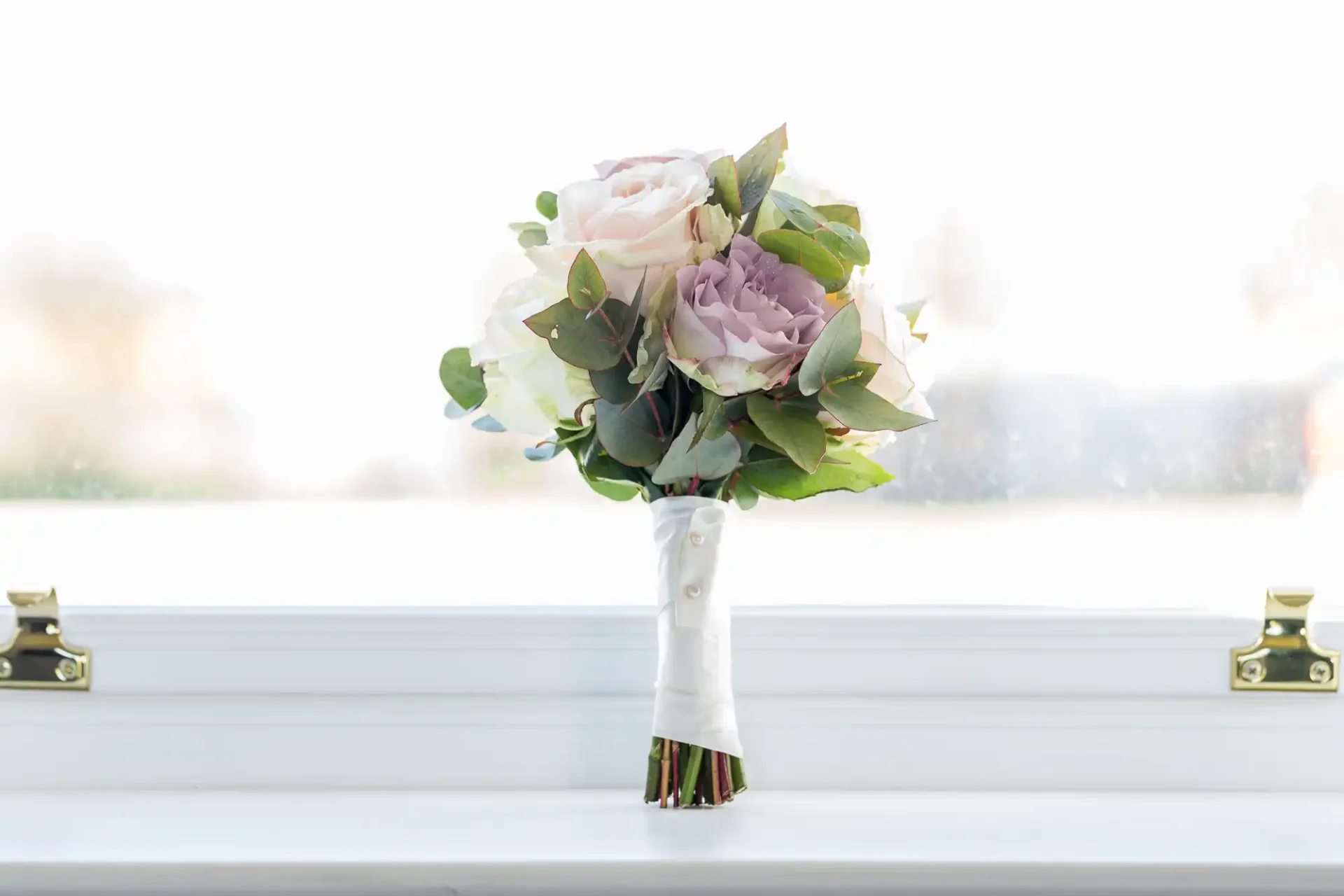 A bouquet of pale purple roses and green foliage in a white vase, placed on a windowsill with a bright background.