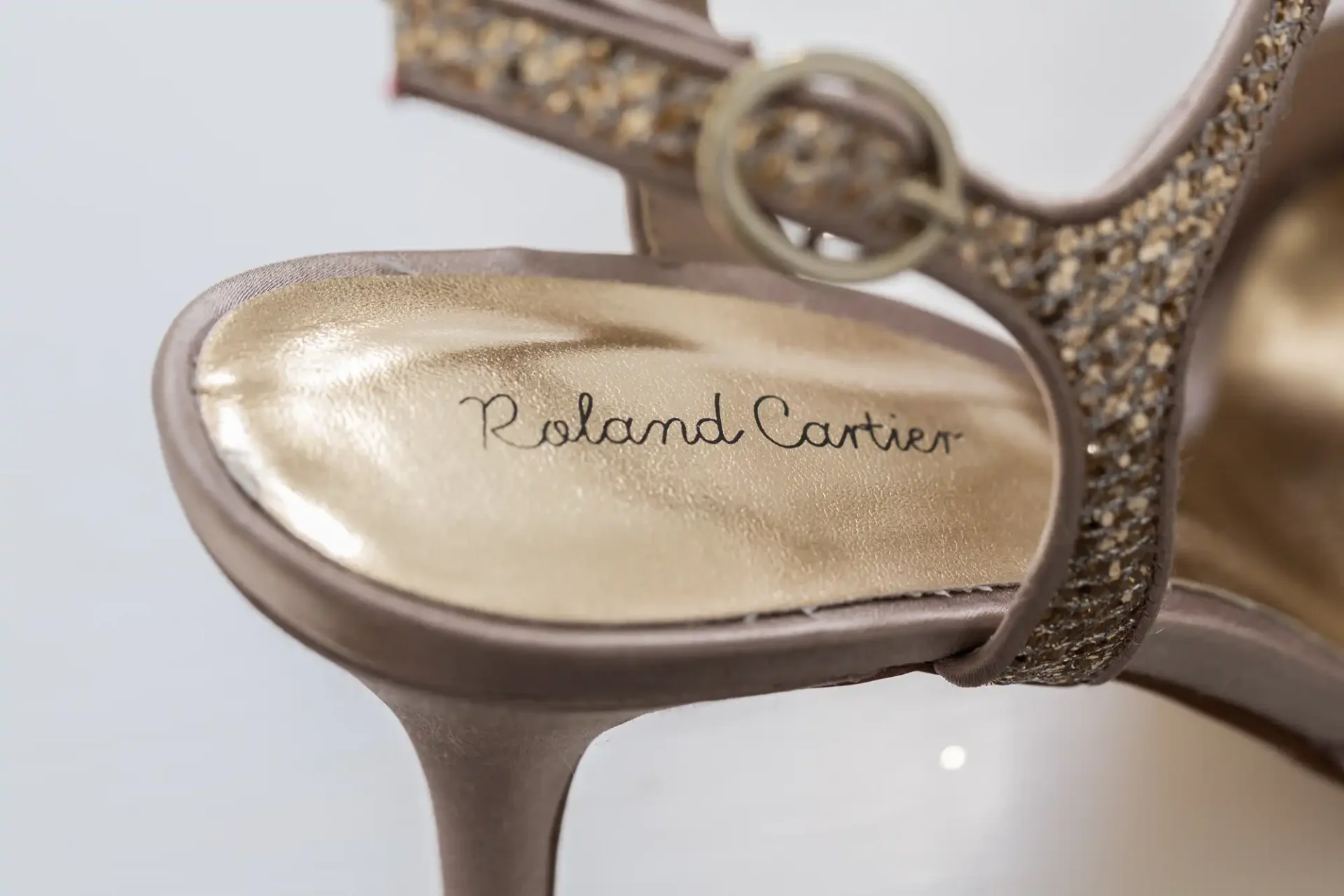 Close-up of a glittery gold high-heeled shoe by roland cartier, focusing on the brand name imprinted on the insole.