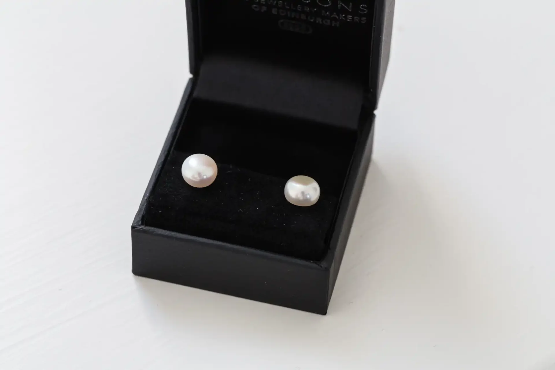 A pair of pearl earrings in an open black jewelry box on a white surface.