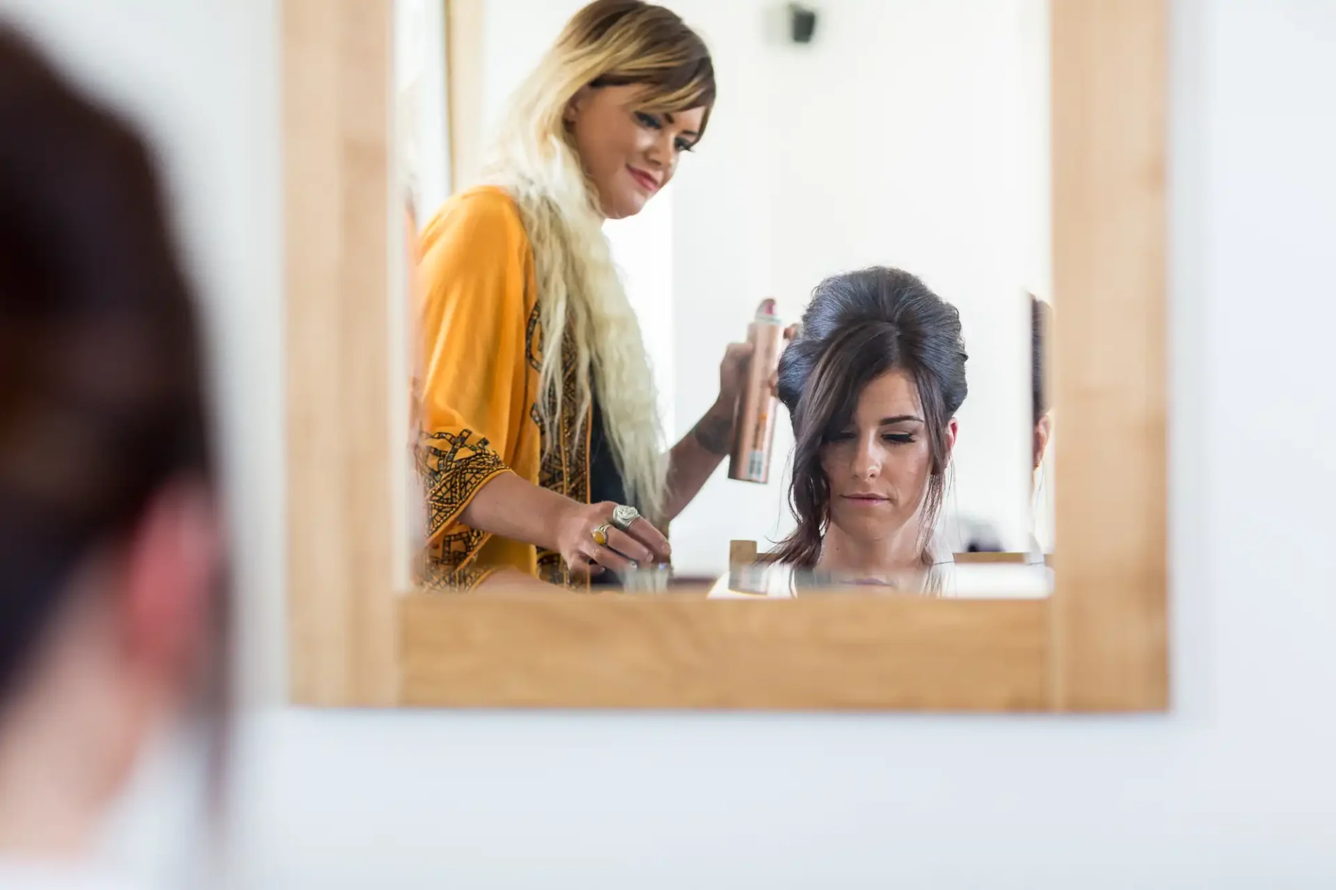 A hairstylist sprays a product on a woman's hair, both reflected in a mirror in a brightly lit salon.
