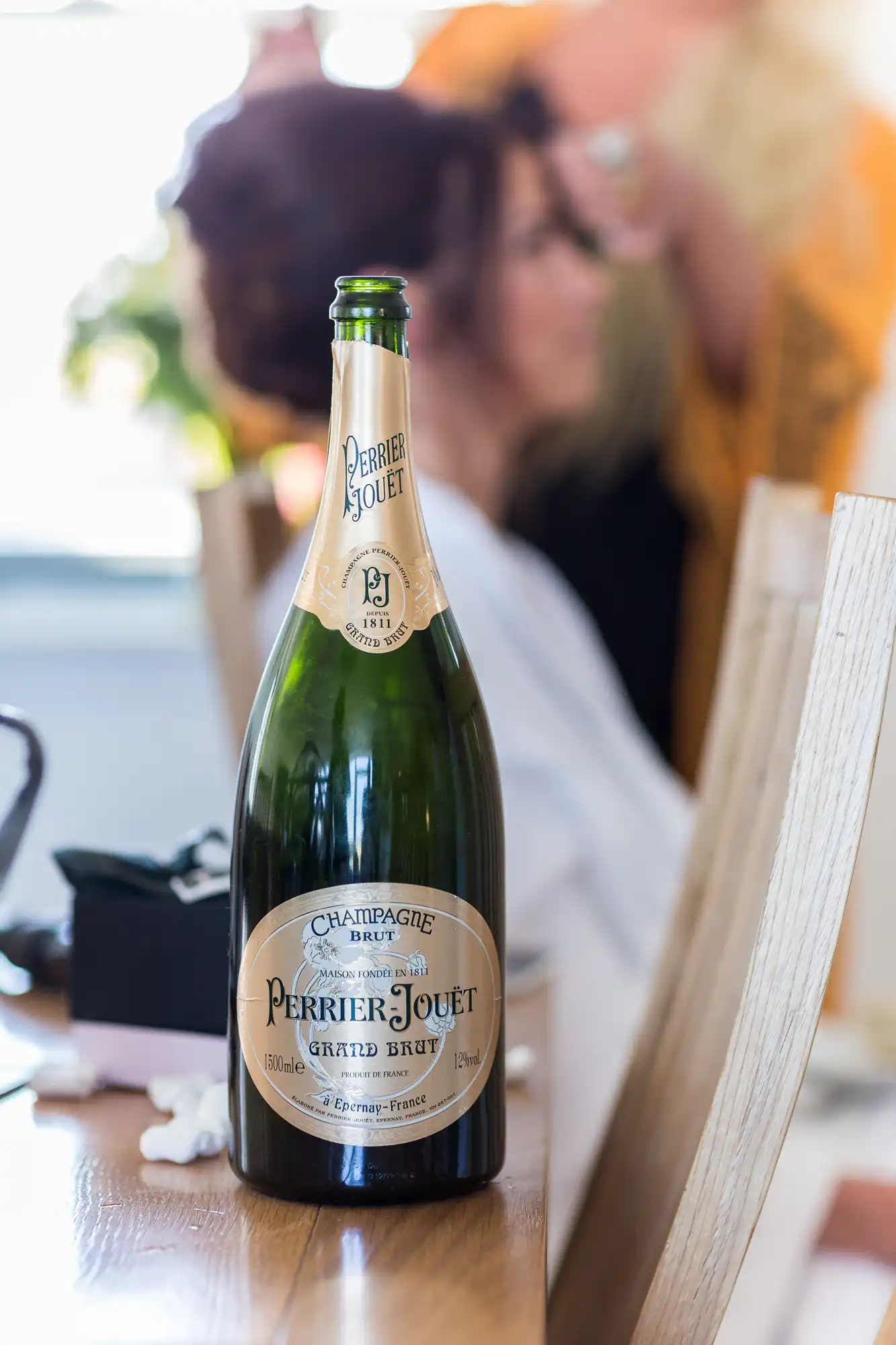 A close-up of a perrier-jouët champagne bottle on a table, with people softly blurred in the background.