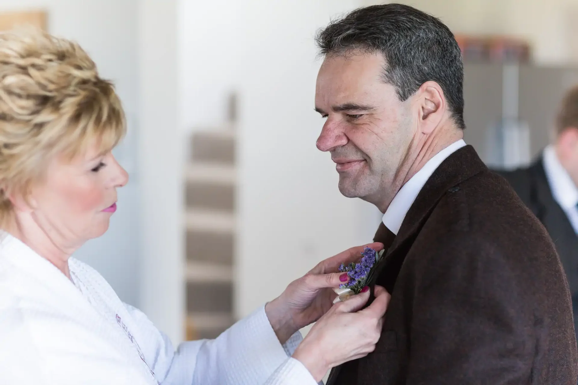 A woman adjusts a boutonniere on a smiling man's lapel in an indoor setting.