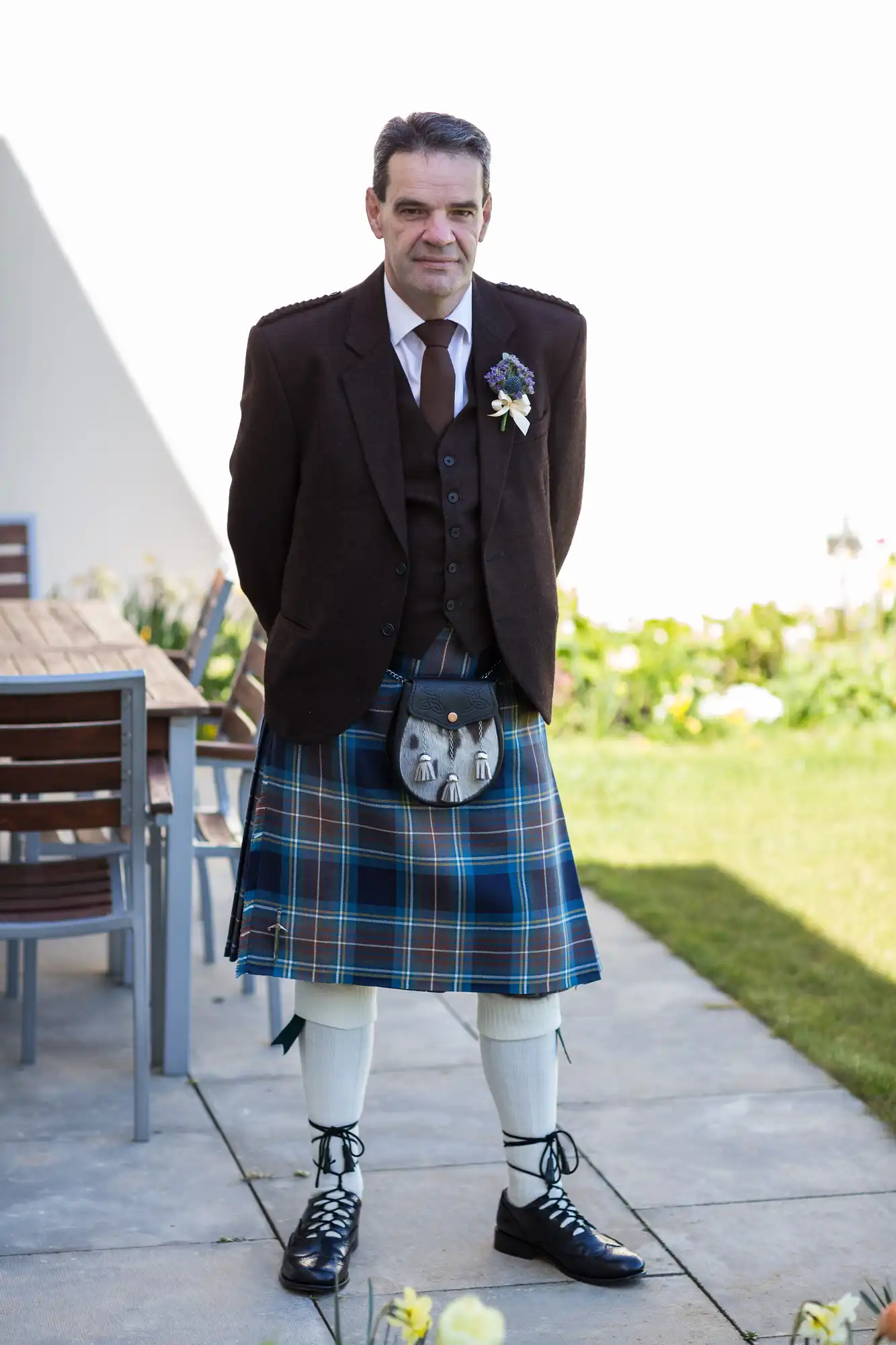 Man standing in a garden dressed in traditional scottish attire, including a tartan kilt, tweed jacket, and sporran.