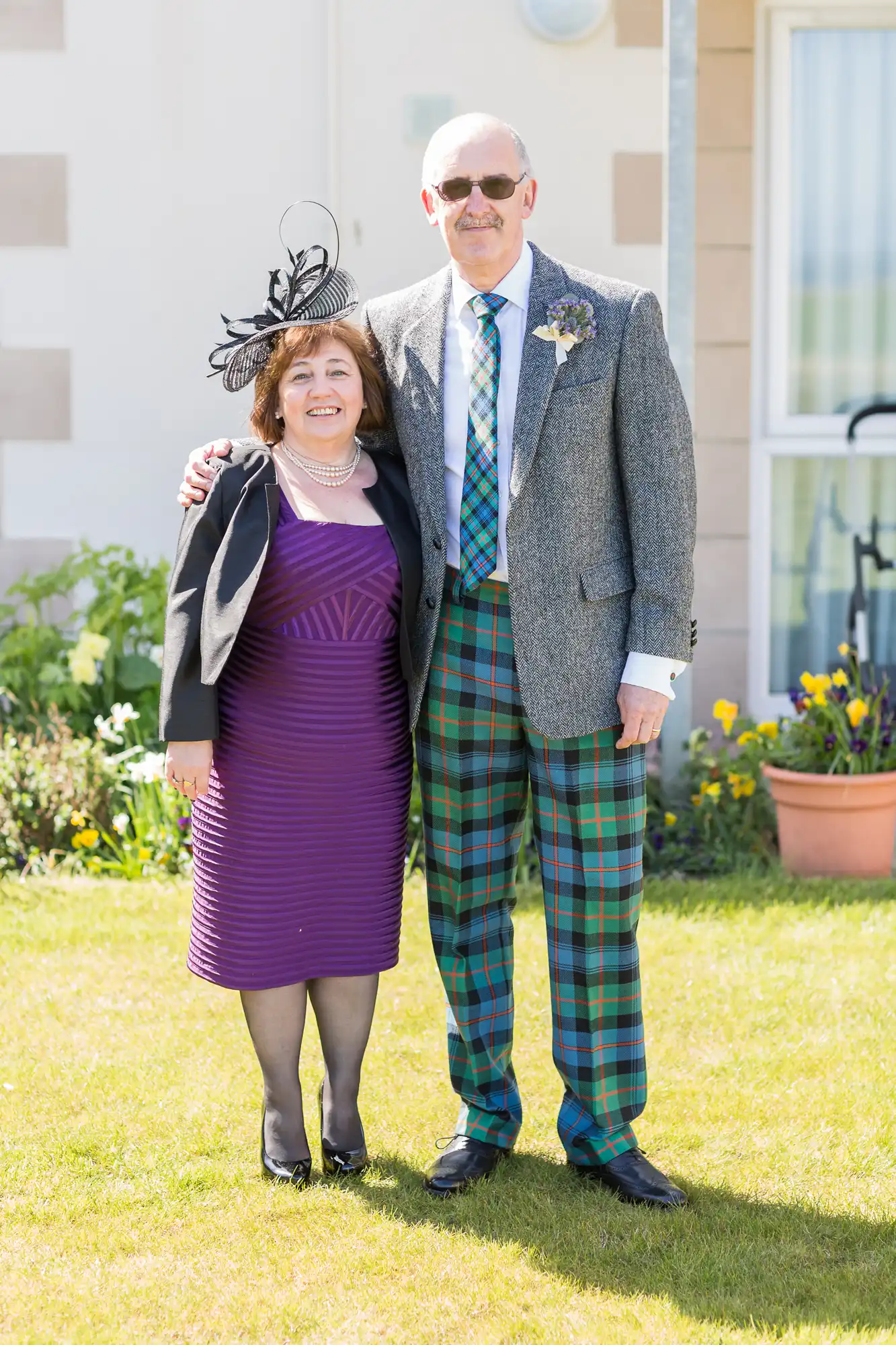 A couple dressed in formal attire, with the man in a tartan kilt and tweed jacket, and the woman in a purple dress and black fascinator, standing together outside on a sunny day.