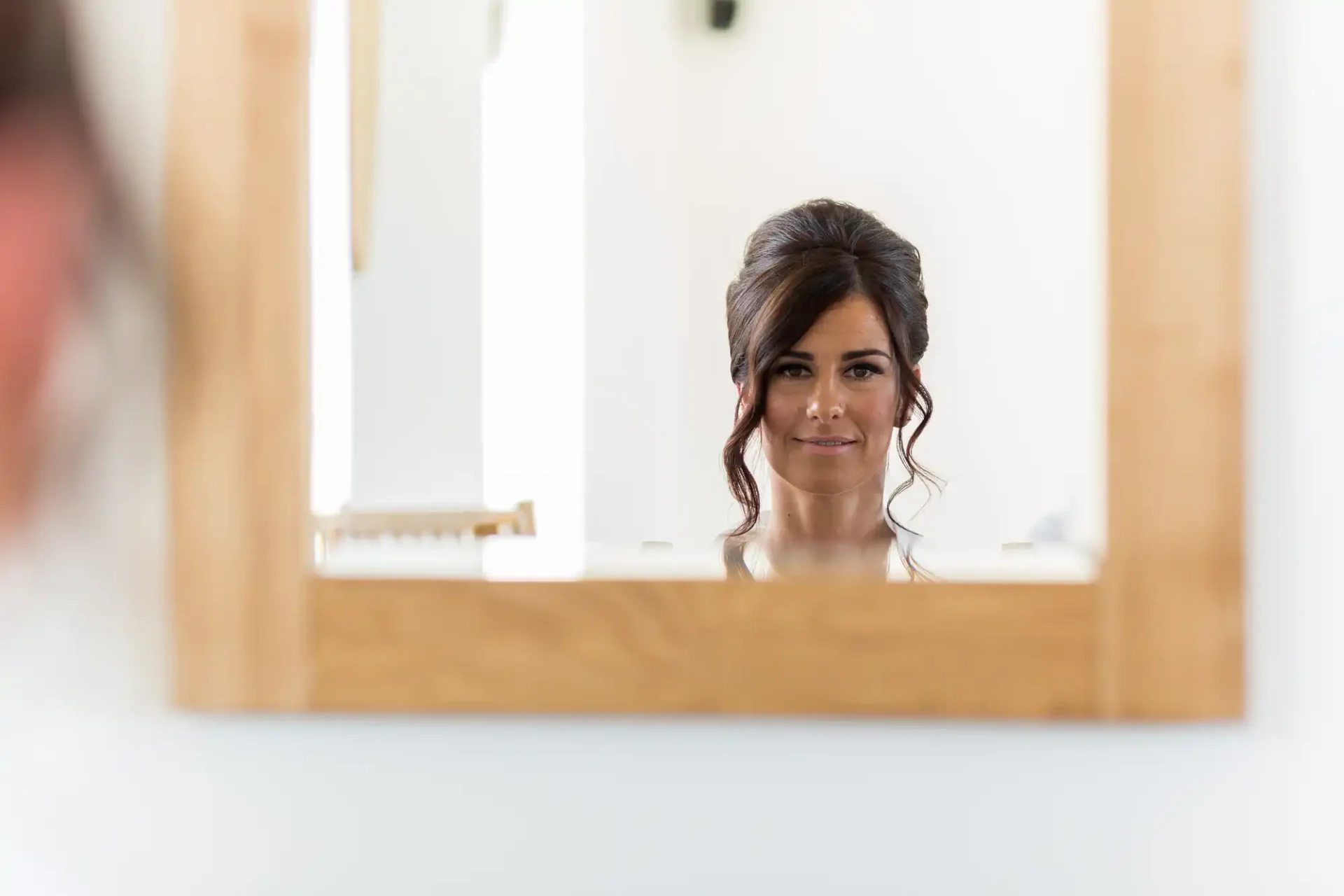 Woman with an updo hairstyle smiling gently, reflecting in a frameless mirror in a bright room.