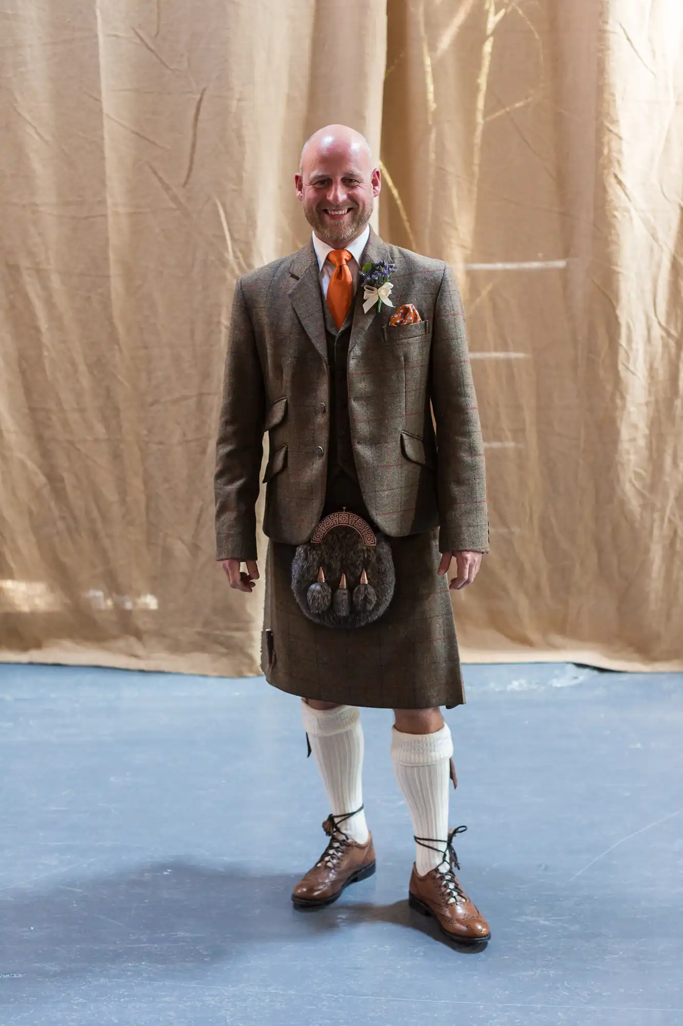 A smiling bald man wearing a traditional scottish outfit with a tweed jacket, kilt, sporran, knee-high socks, and brown shoes, standing against a beige backdrop.