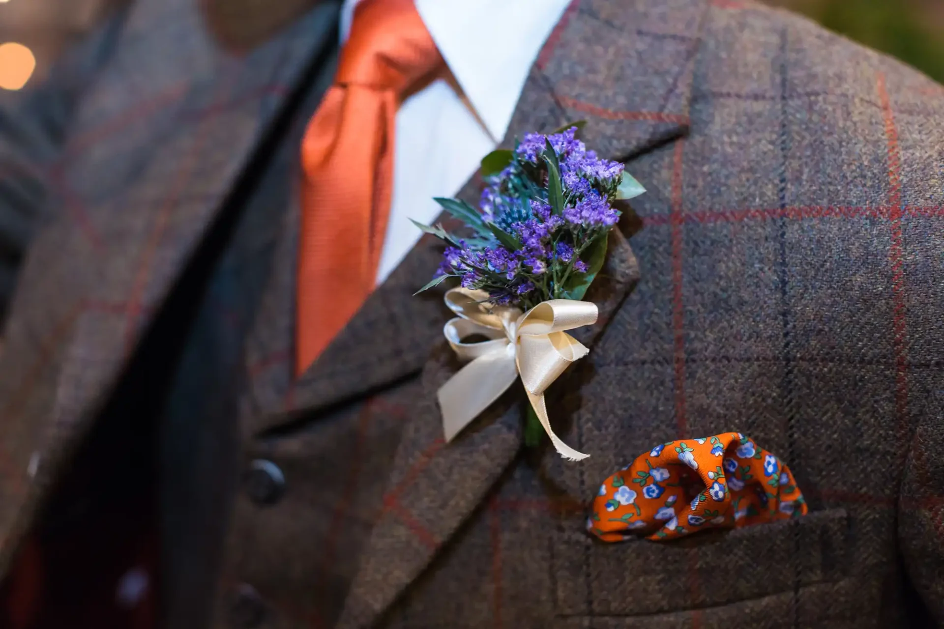 A close-up of a man's chest wearing a tweed suit with a purple patterned pocket square and a boutonniere featuring purple flowers and a cream ribbon.
