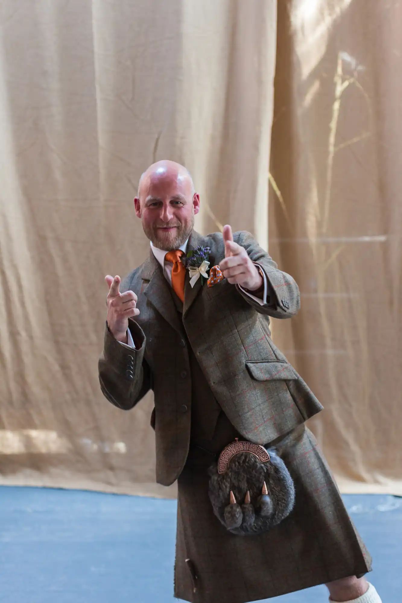 A bald man in a tweed suit and kilt, smiling and gesturing thumb's up with both hands, stands in front of a beige curtain.