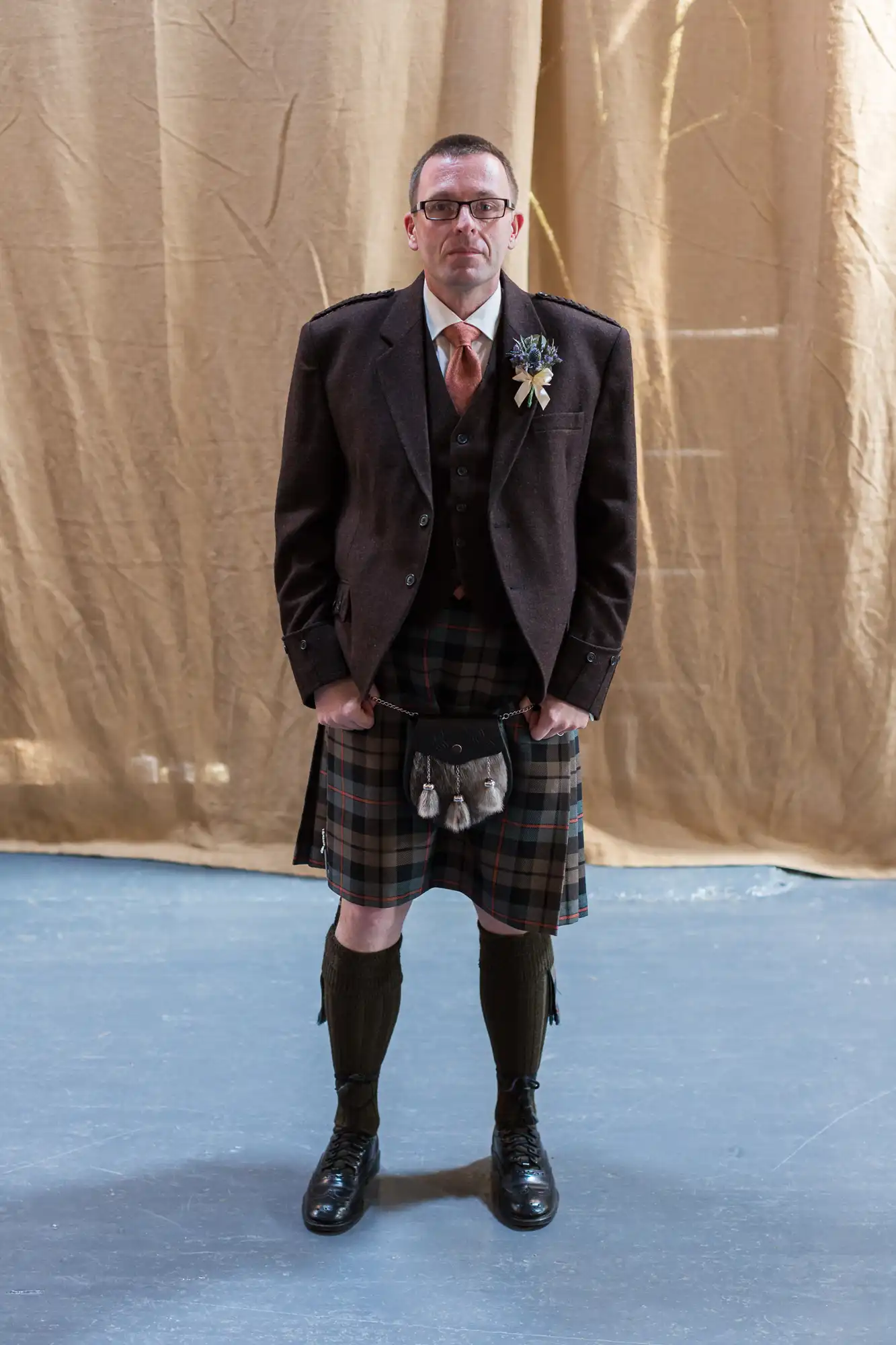 Man in traditional scottish attire including a tweed jacket, kilt, sporran, and knee-high socks, standing against a beige backdrop.