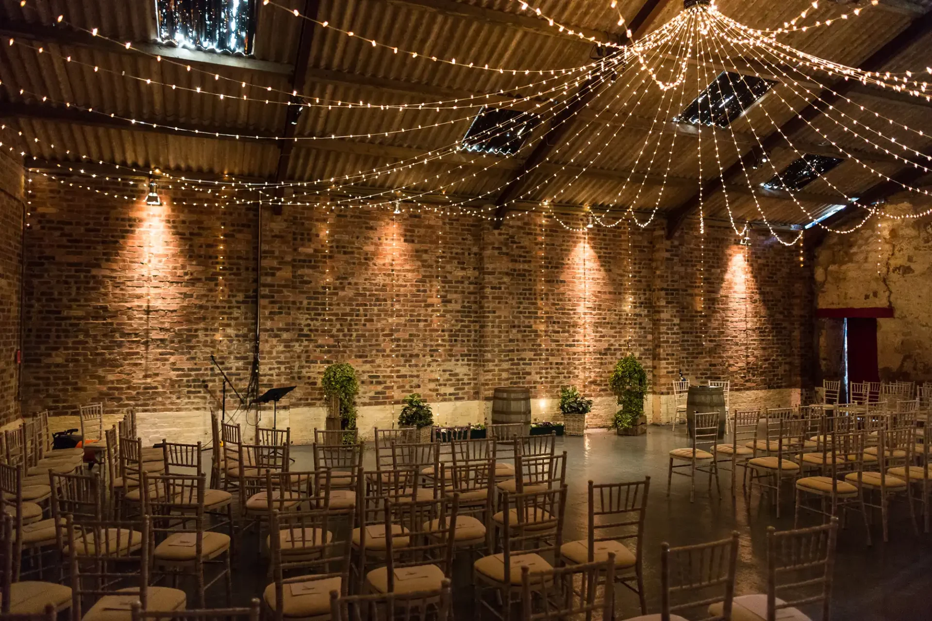 Rustic indoor venue with brick walls and wooden beams, adorned with string lights and set up with rows of chairs for an event.