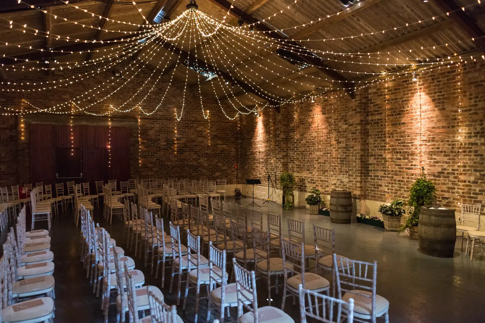 Rustic event space with exposed brick walls, draped string lights and arranged white chairs facing a central aisle.