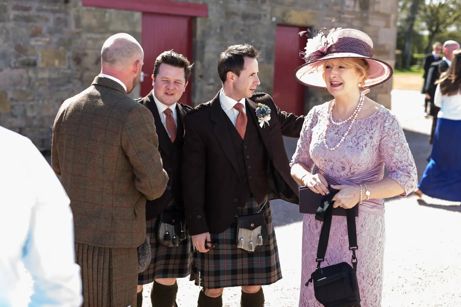 Group of people in formal attire including kilts, conversing outdoors at a sunny event, with a woman in a hat smiling broadly.