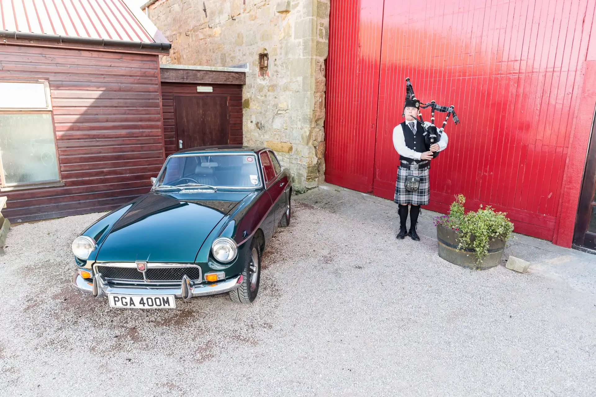 A man in traditional scottish attire playing bagpipes beside a classic dark blue mg car, in a courtyard with red and stone walls.