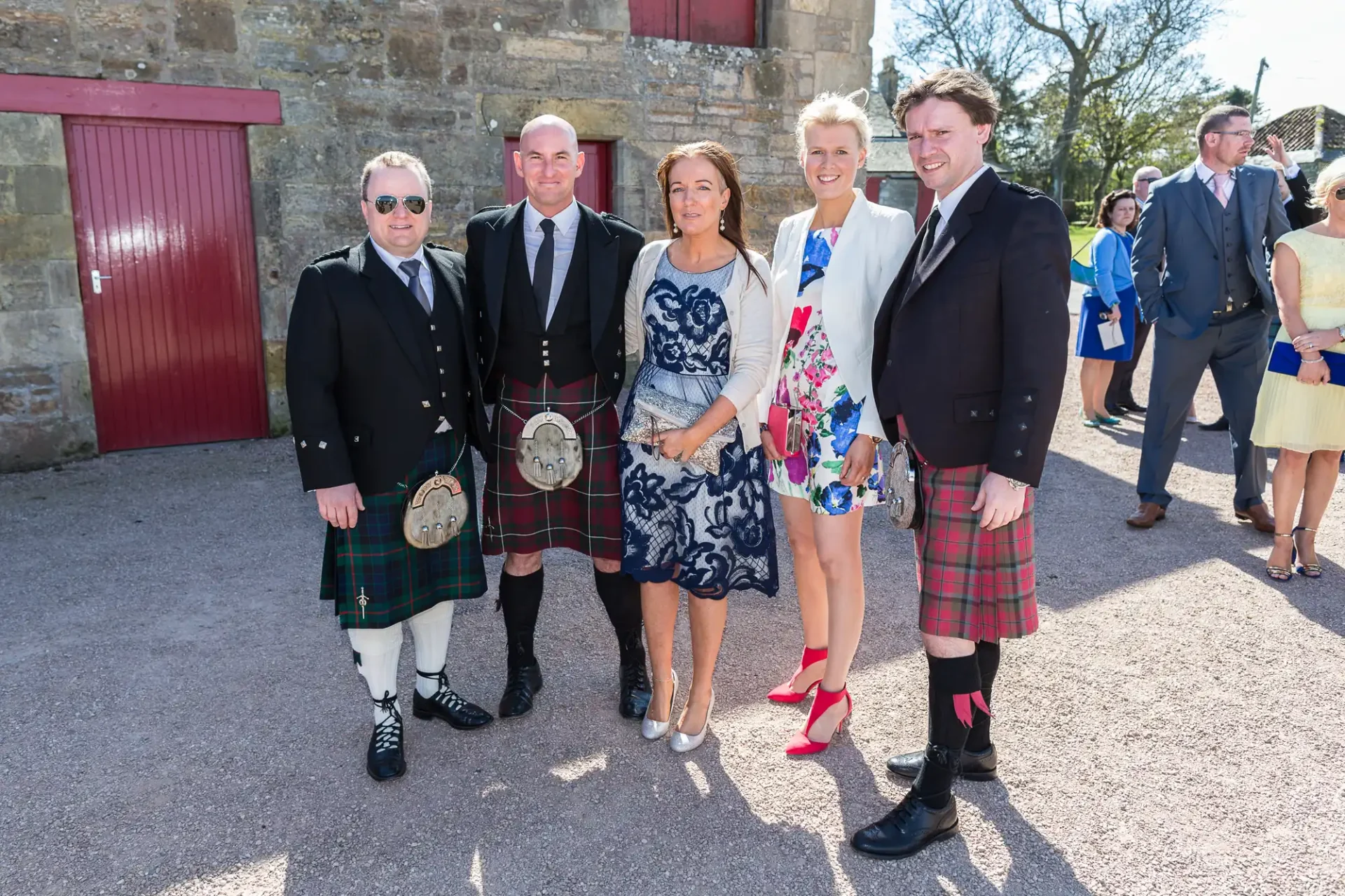 Group of five adults in formal attire, including kilts and dresses, standing outside at a social event on a sunny day.