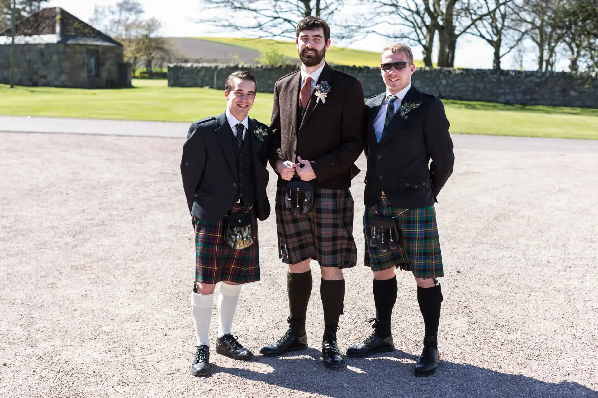 Three men in traditional scottish kilts and jackets posing together outdoors on a sunny day, with a stone wall and trees in the background.