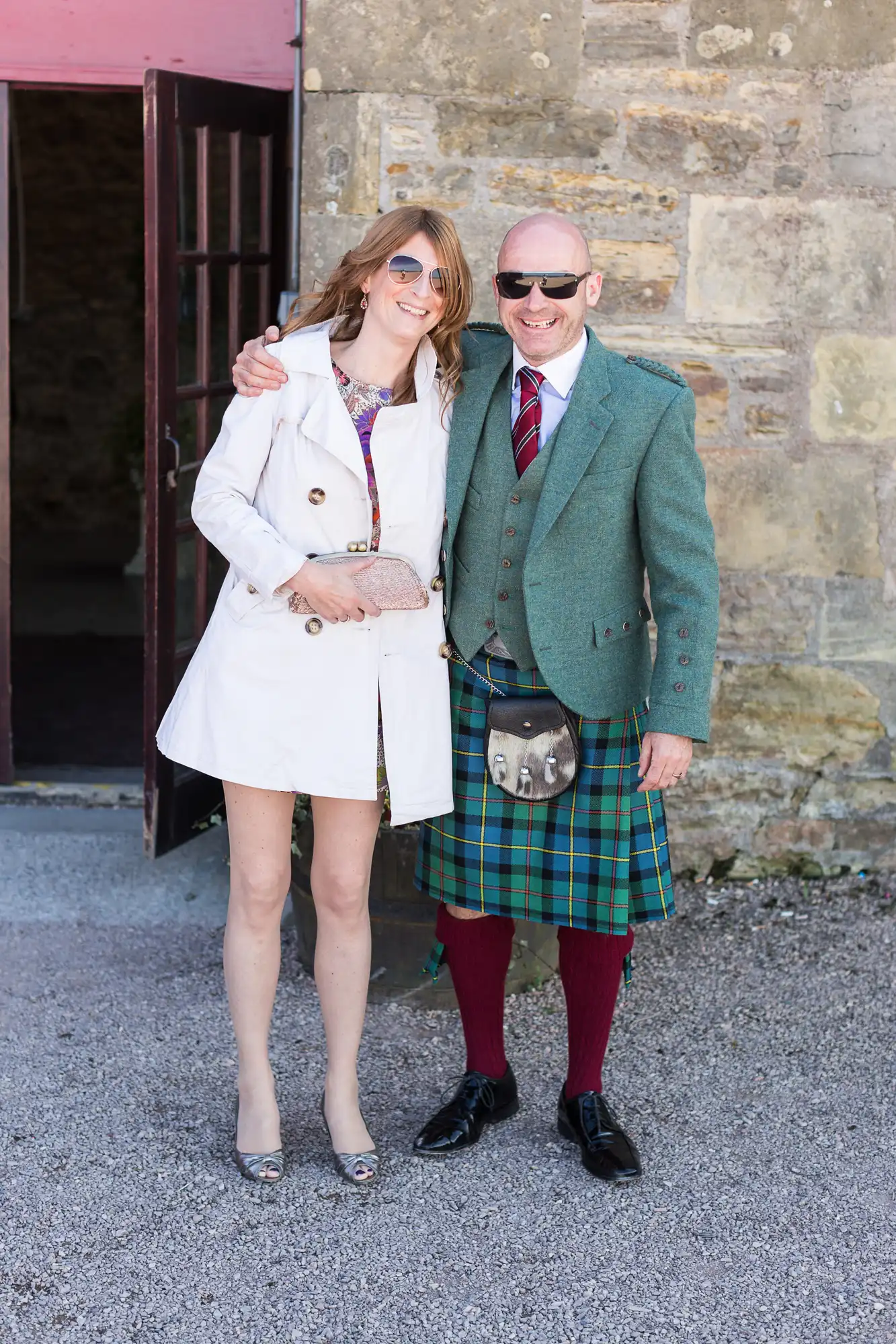 A woman in a white trench coat and a man in a green blazer and kilt smiling arm-in-arm outside a stone building.