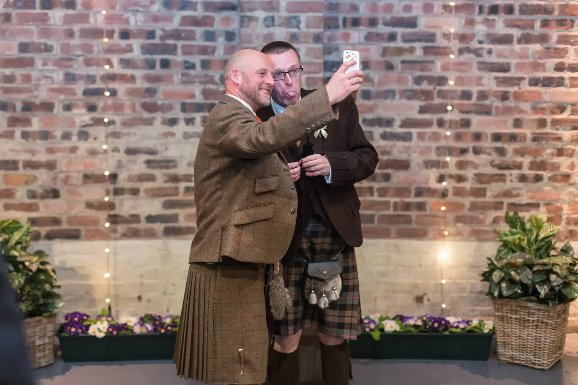 Two men, one in a suit and the other in a kilt, happily taking a selfie together at a venue with brick walls and potted plants.