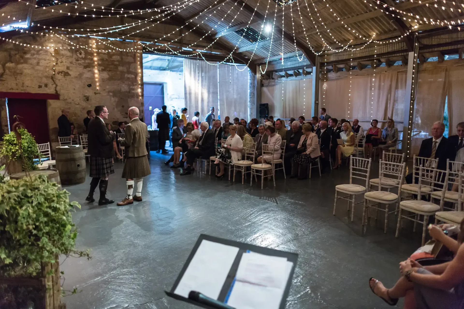 Guests in a rustic event space with string lights, listening to a speaker in traditional scottish attire.