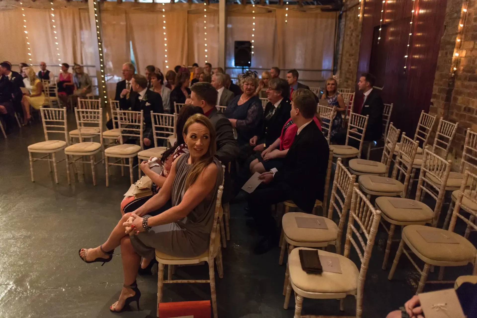 Guests seated on chairs at an indoor wedding venue with string lights, looking towards the front where the ceremony is taking place.