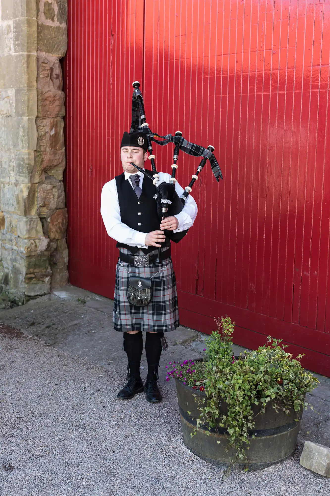 A man in traditional scottish attire, including a kilt and sporran, plays bagpipes beside a red wall and a planter with flowers.