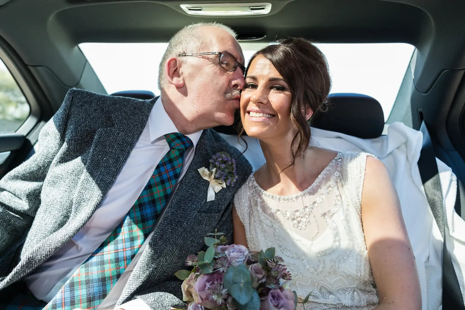 An elderly man in a tartan vest kisses a smiling young woman in a wedding dress, both seated in a car, holding a bouquet.