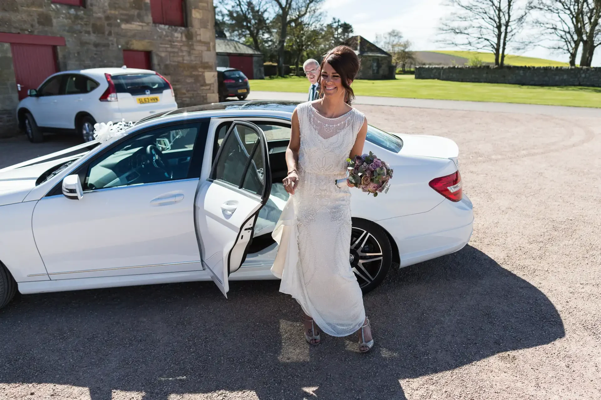 A bride in a white dress holding a bouquet steps out from a white luxury car on a sunny day.